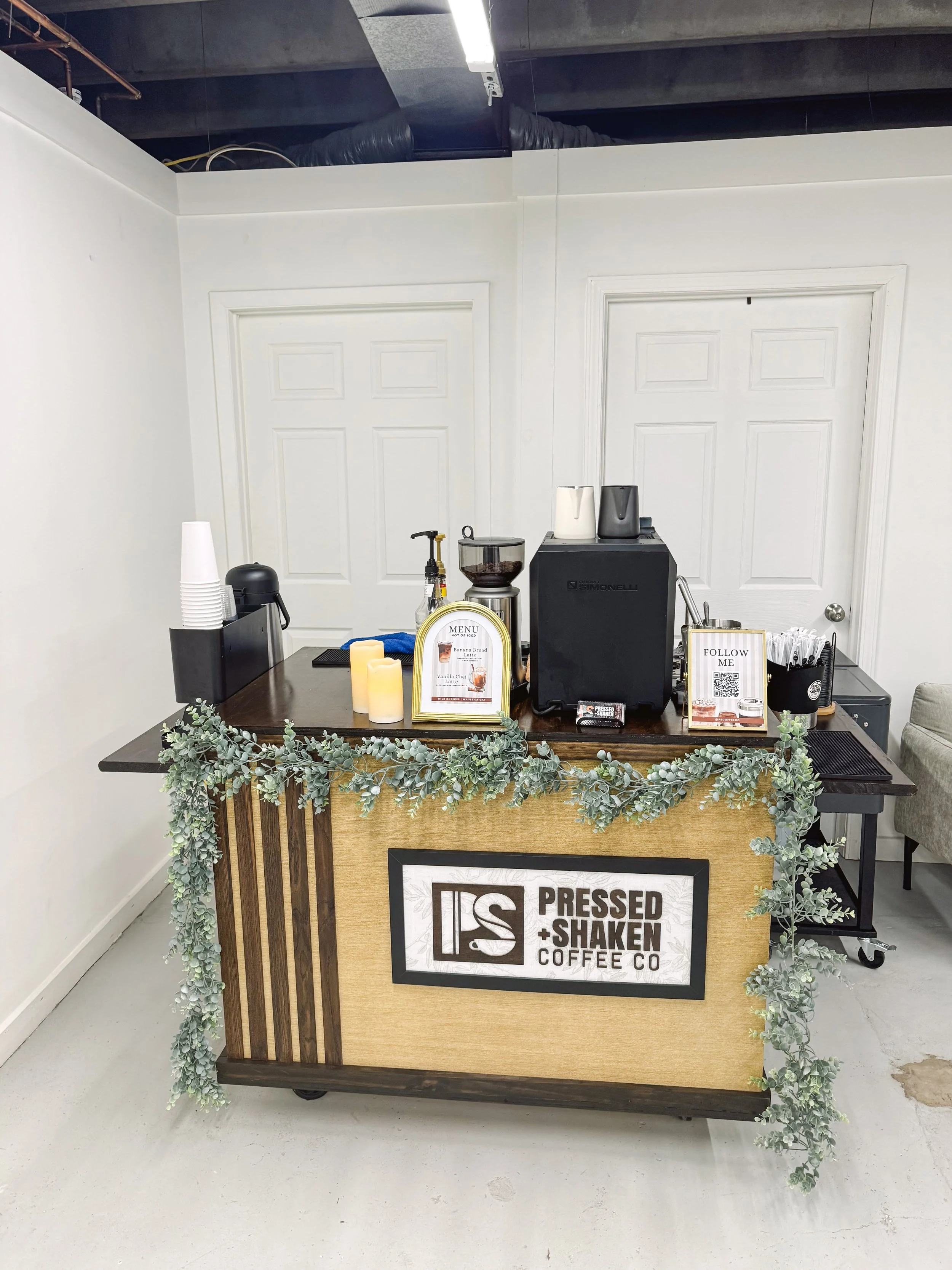 Coffee stand with a sign reading 'Pressed + Shaken Coffee Co', decorated with greenery and candles, featuring a coffee maker and cups.