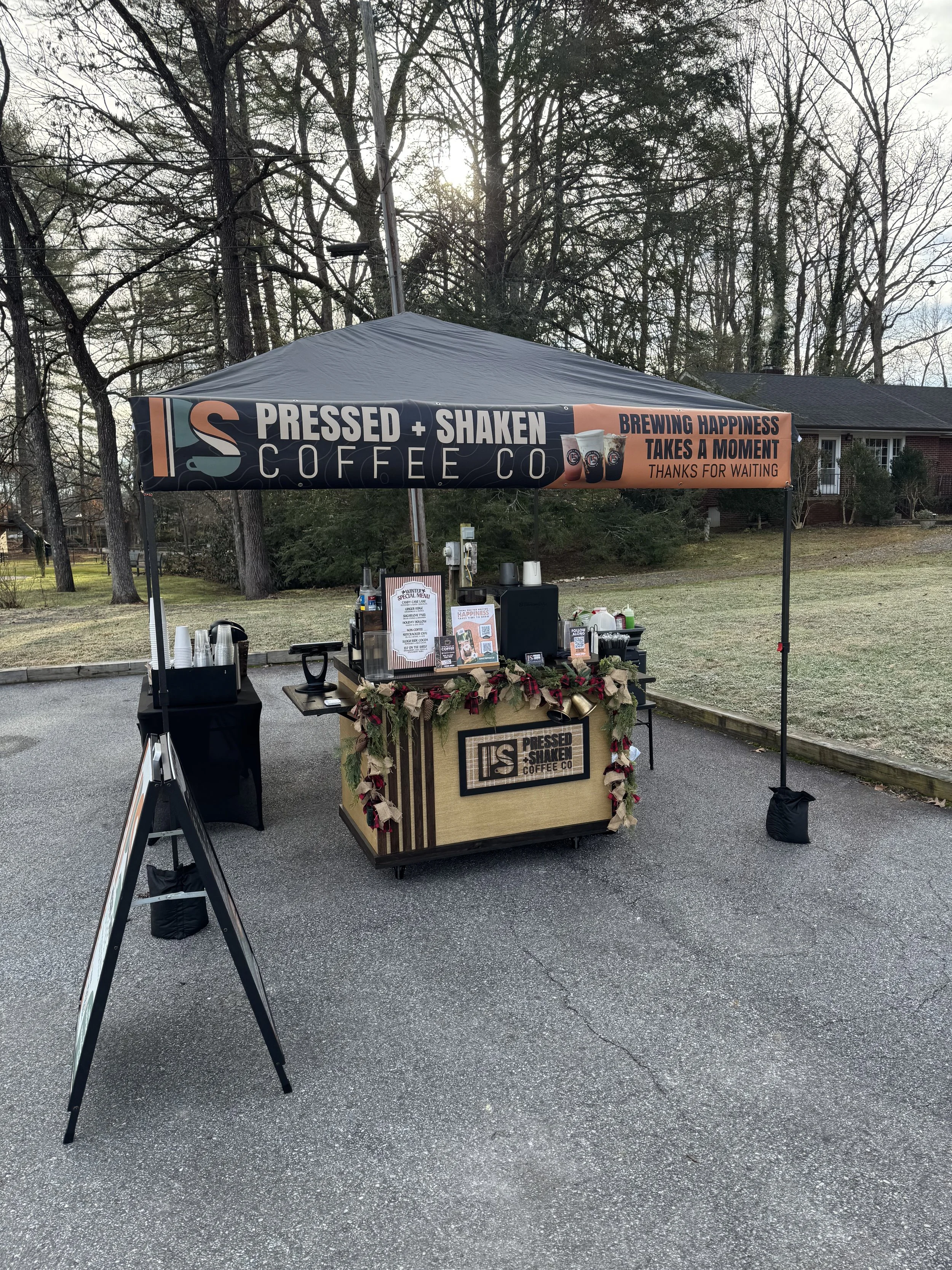 Outdoor coffee stand with a black tent from Pressed + Shaken Coffee Co., decorated with holiday ornaments, set up on pavement with trees and a house in the background, serving coffee and beverages.