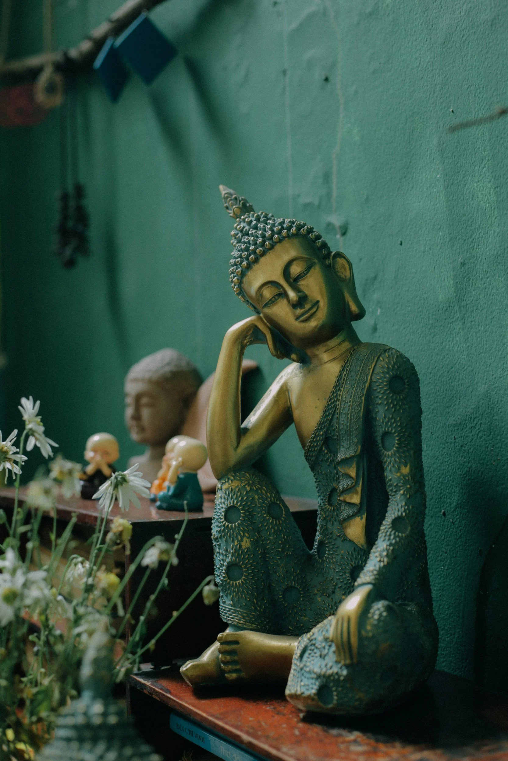 A decorative Buddha statue sitting with one arm resting on its knee and head leaning on its hand, surrounded by smaller figurines and flowers against a green wall.