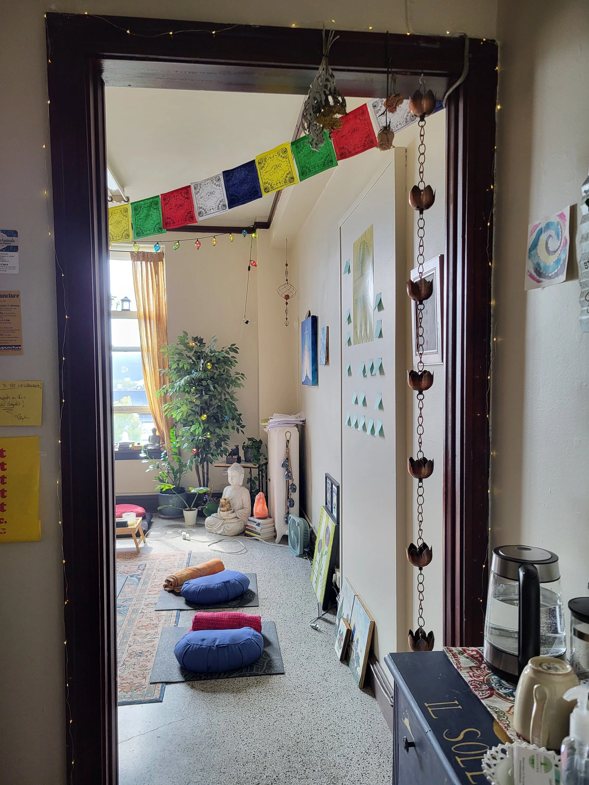 Room with cross-legged meditation cushions, a statue of Buddha, potted plants, wall art, and a window with orange curtains, decorated with colorful prayer flags and string lights.