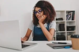 Young woman with curly hair and glasses working on a laptop at a desk in a home office.