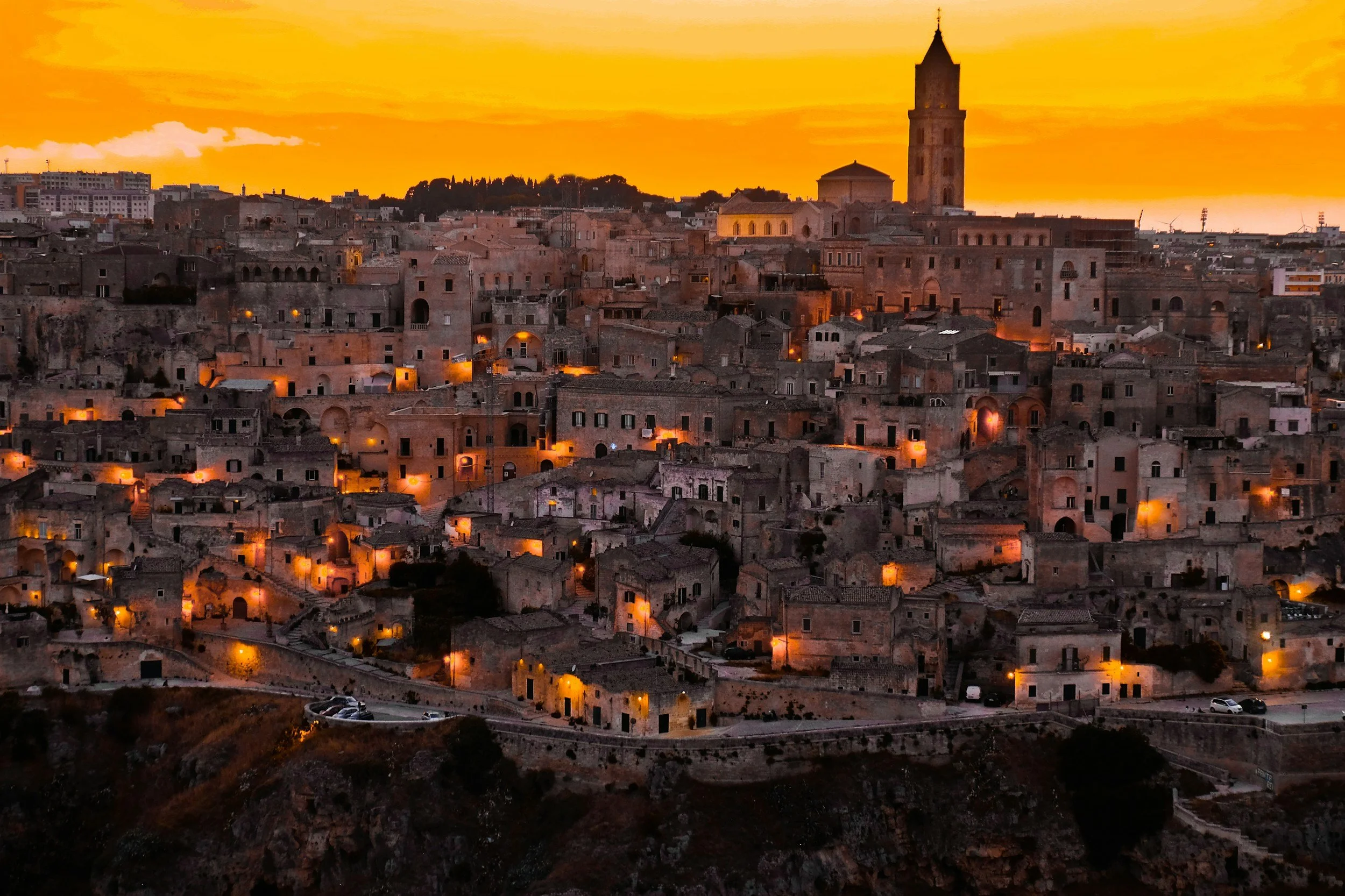Sunset over an ancient city with stone buildings, narrow streets, and a tall church tower in the distance.