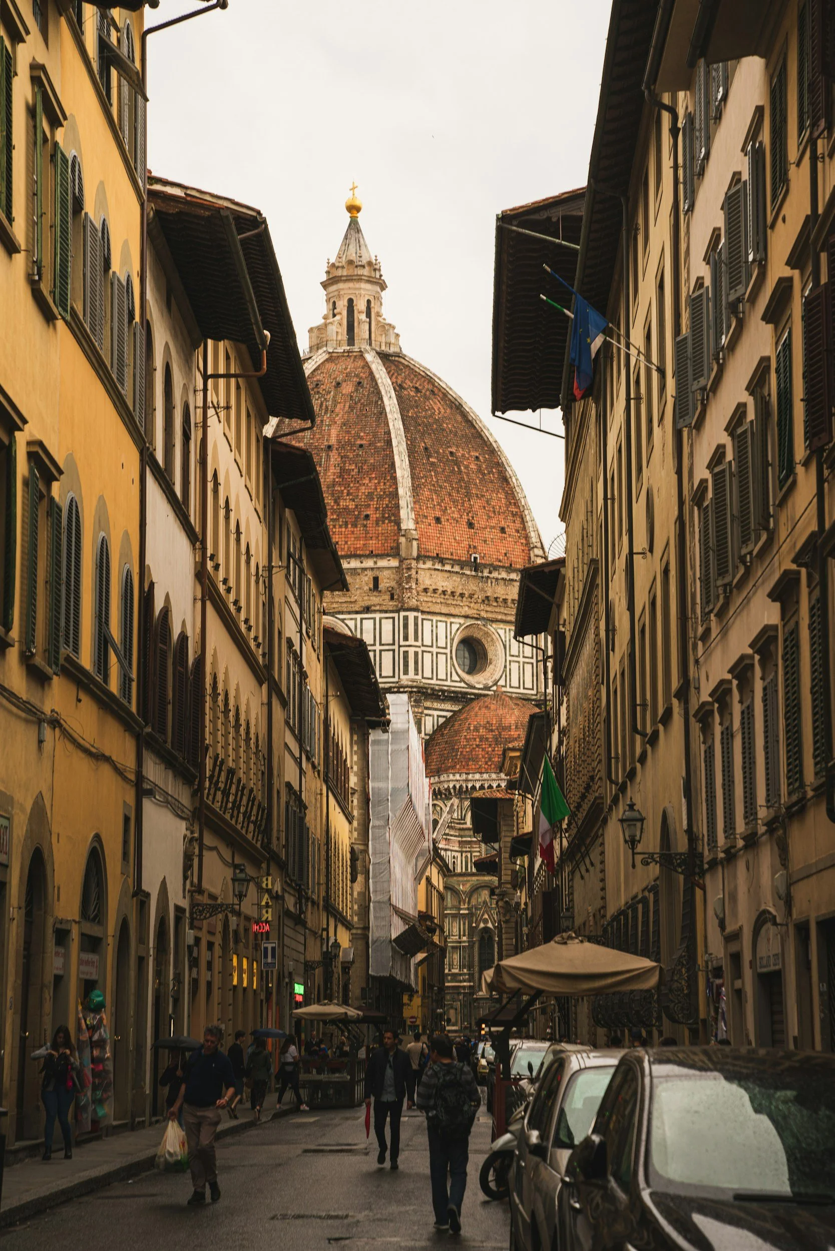 A narrow street in Florence, Italy, with yellow and cream buildings, some with Italian flags, and the iconic Florence Cathedral (Duomo) with its large red-tiled dome in the background.