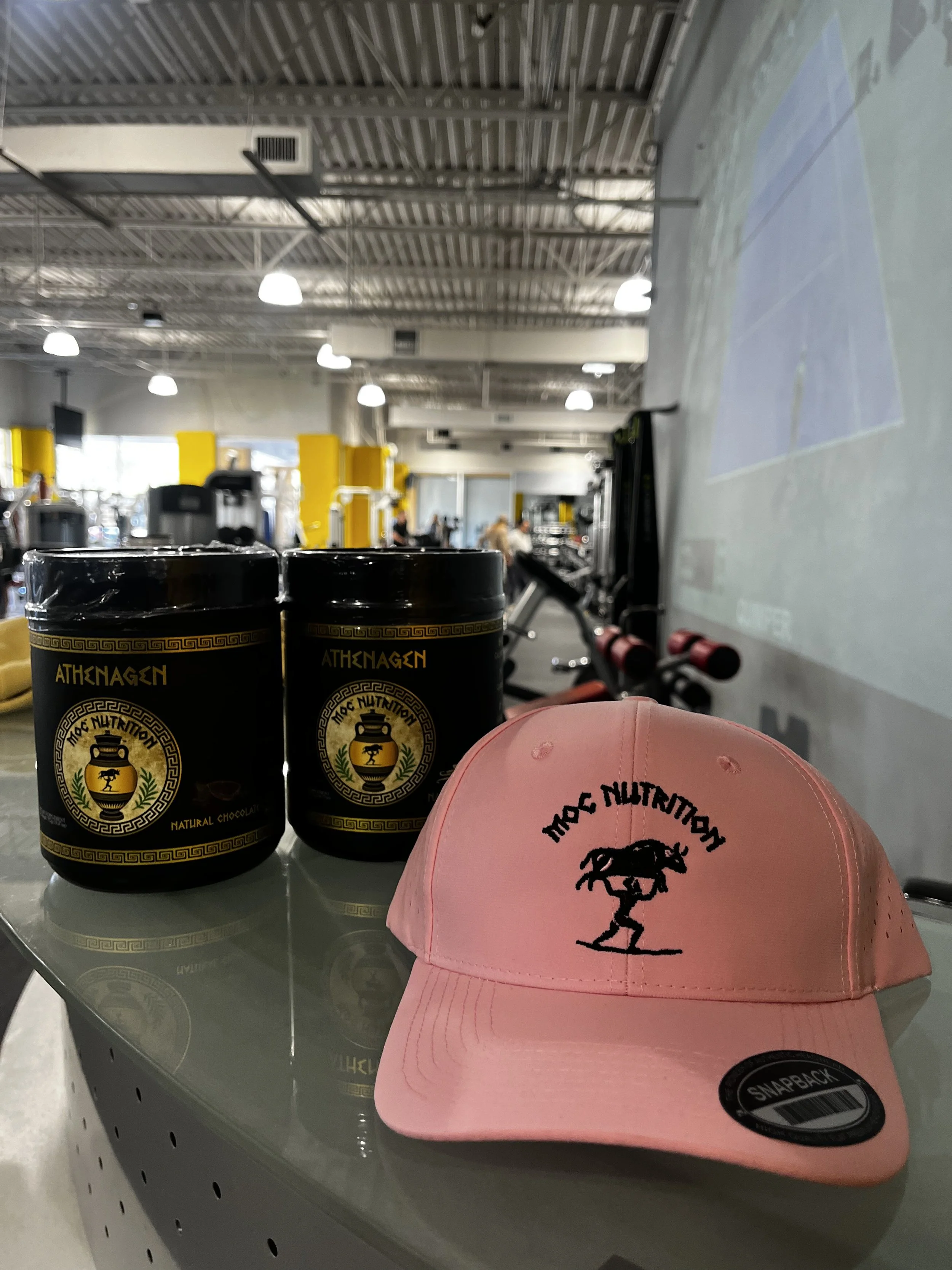 Two black containers of MOC Nutrition supplements, a pink hat with the logo of MOC Nutrition, and gym equipment in the background of a gym interior.