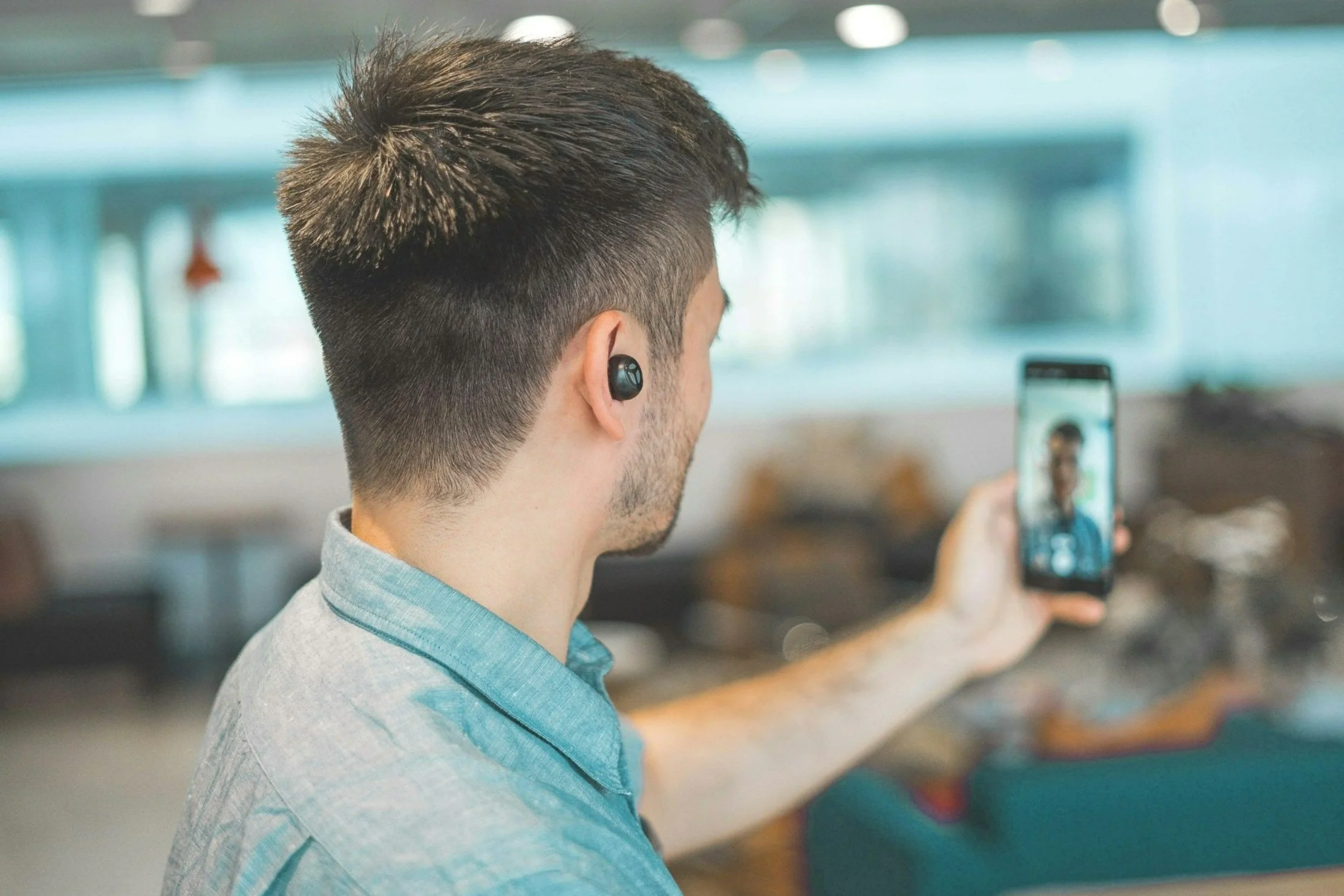 A man taking a selfie with his smartphone in a well-lit indoor space, wearing wireless earbuds and a casual light blue shirt.