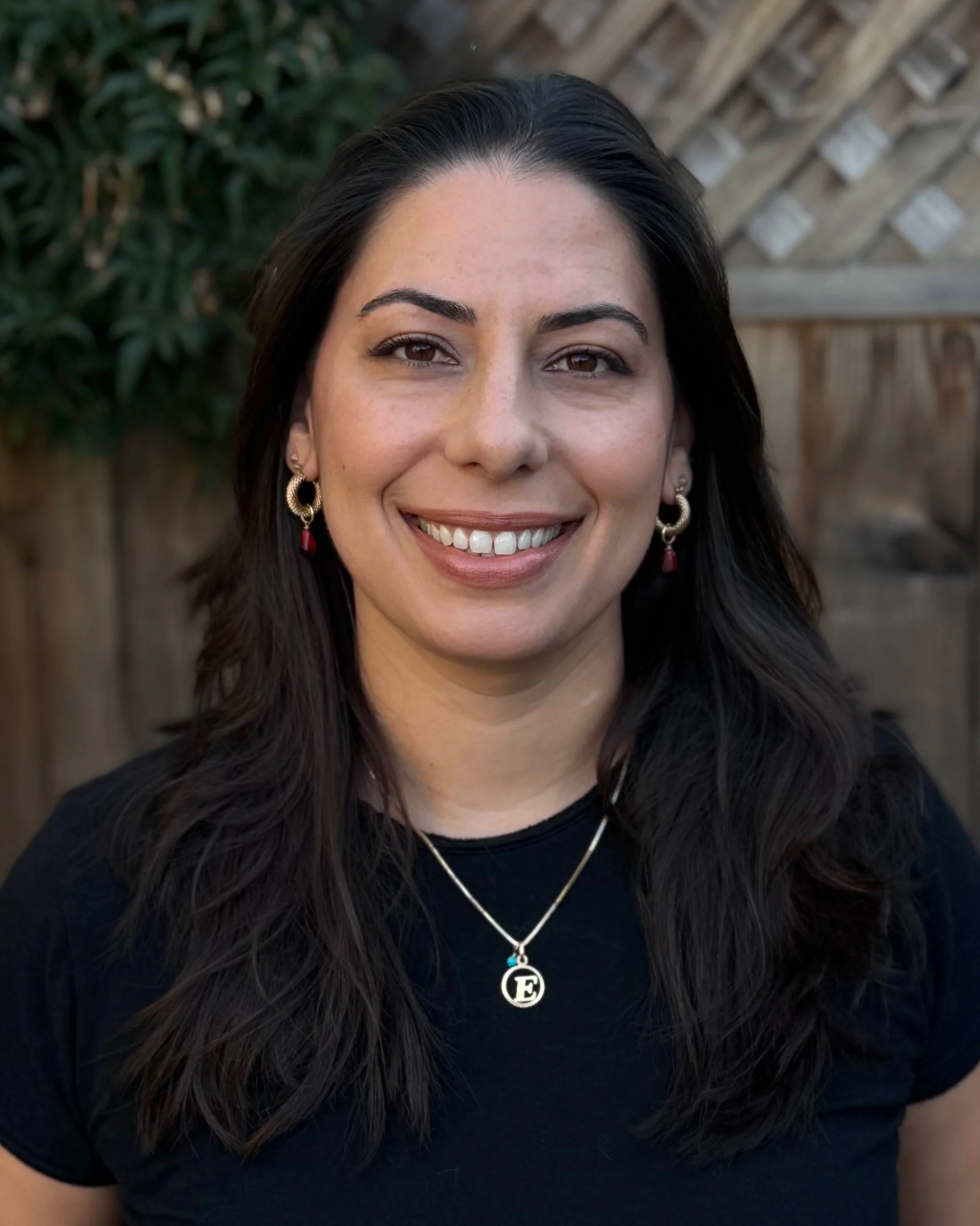 Close-up of a smiling woman with long dark hair, wearing earrings, a necklace with a pendant that has the letter E, and a black top. Background includes a wooden fence and greenery.