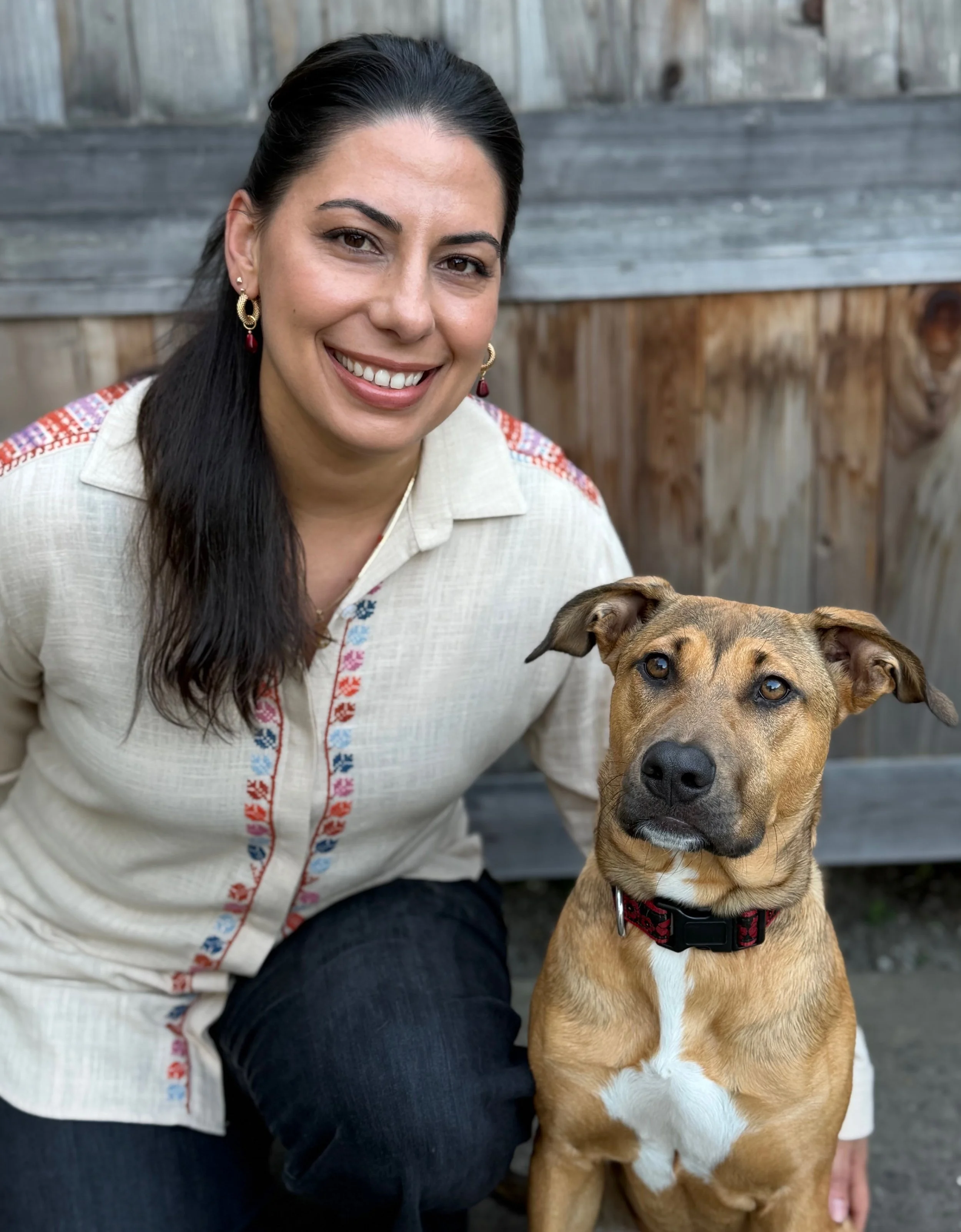 A woman with long dark hair and earrings smiling at the camera, sitting outdoors next to a tan and white dog with a red collar, in front of a wooden fence.