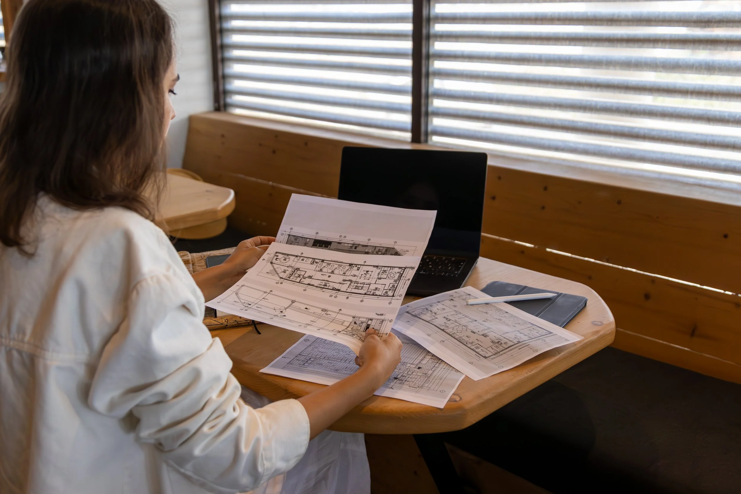 Mujer revisando planos arquitectónicos en una mesa de madera, con una computadora portátil y documentos adicionales en un ambiente con ventanas con persianas.