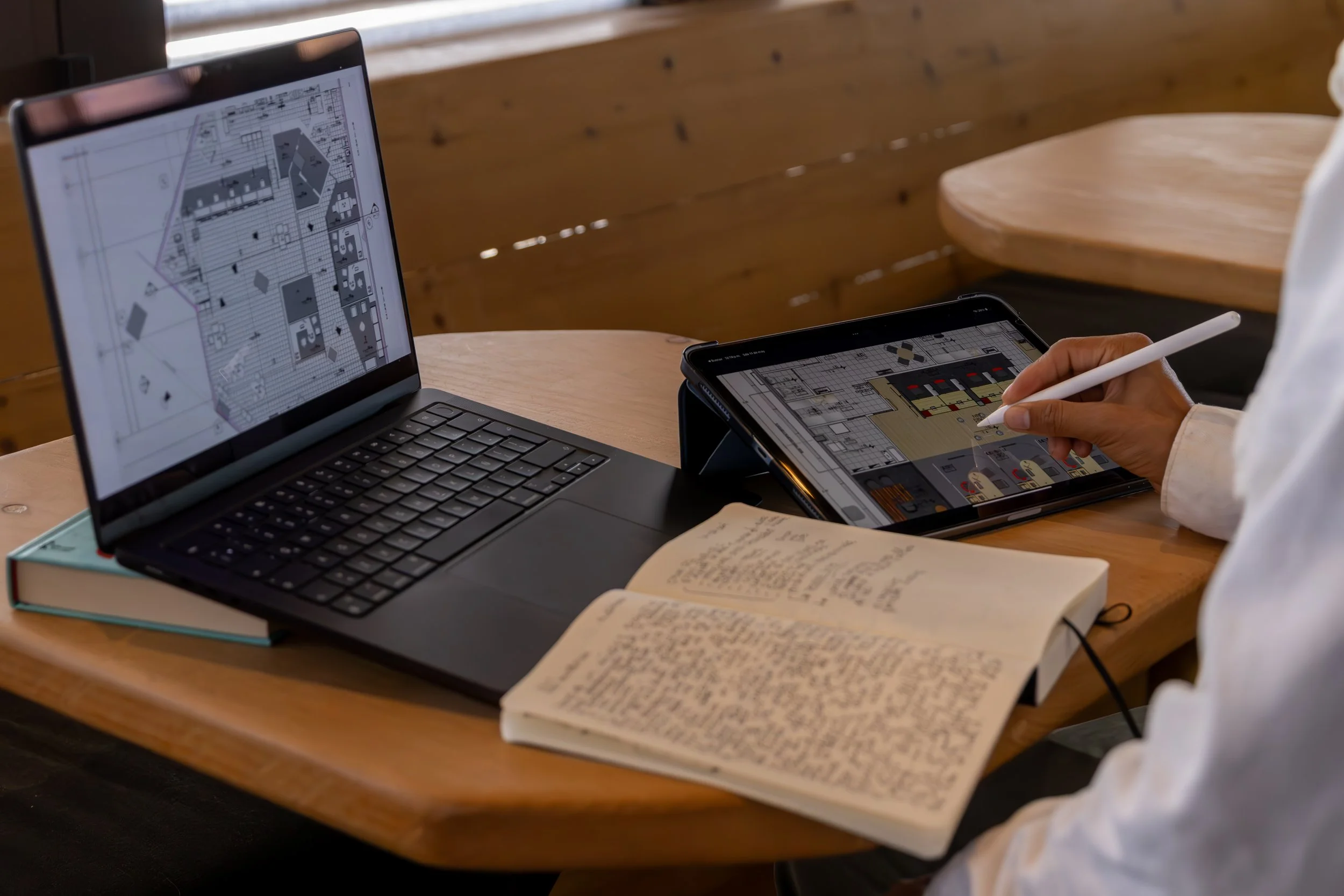 Persona trabajando en diseño de interiores con laptop, tableta y cuaderno en un ambiente de madera.