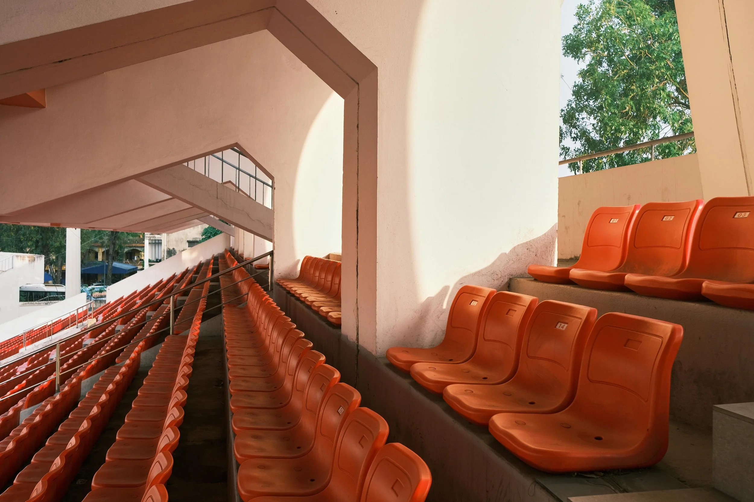 Asientos de estadio de color naranja en un graderío exterior, con sombra y un árbol verde visible al fondo.