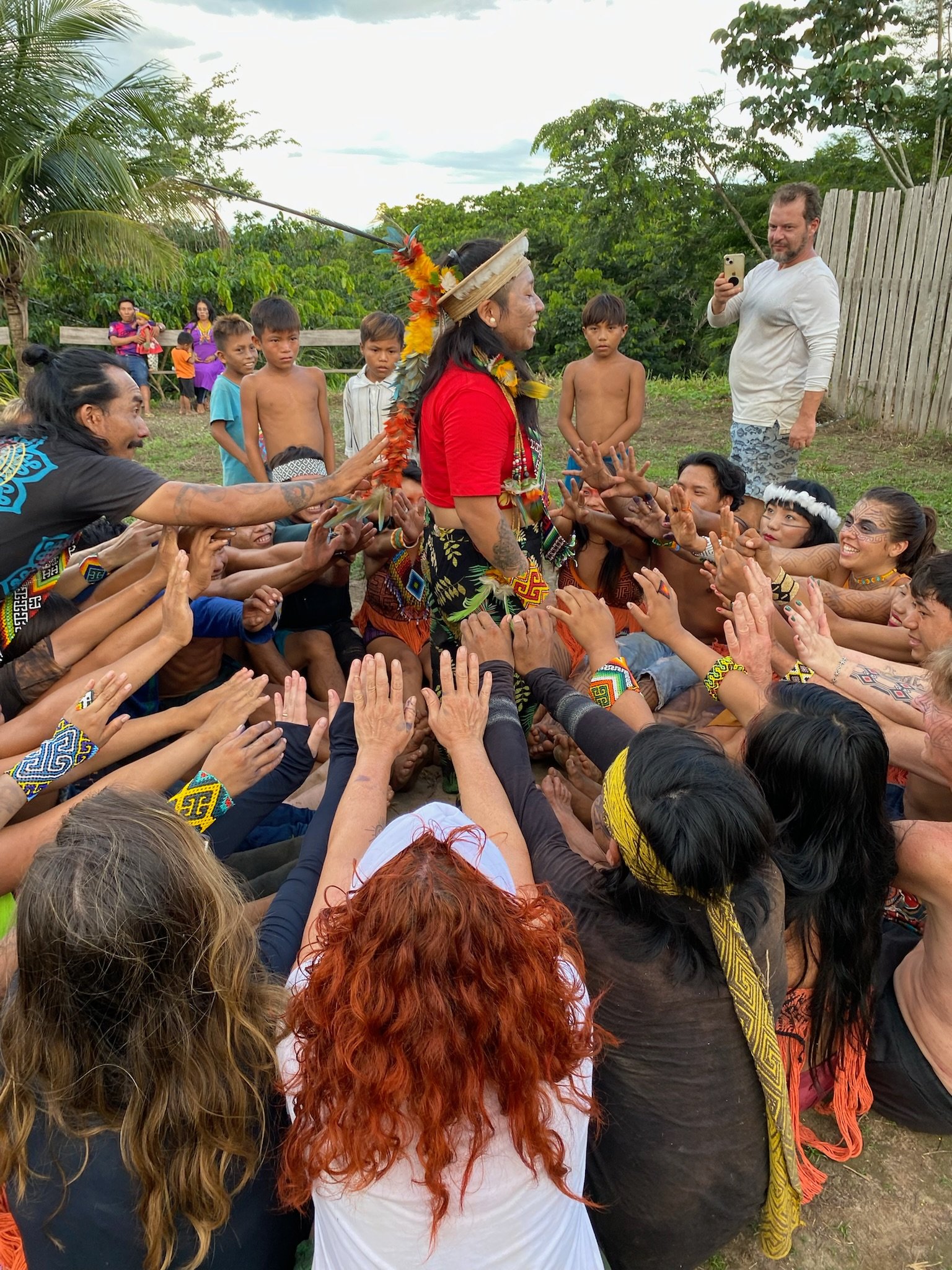 Mulher com roupas tradicionais indígenas, usando adereço de penas, está cercado por pessoas em um ambiente ao ar livre com árvores e céu nublado.