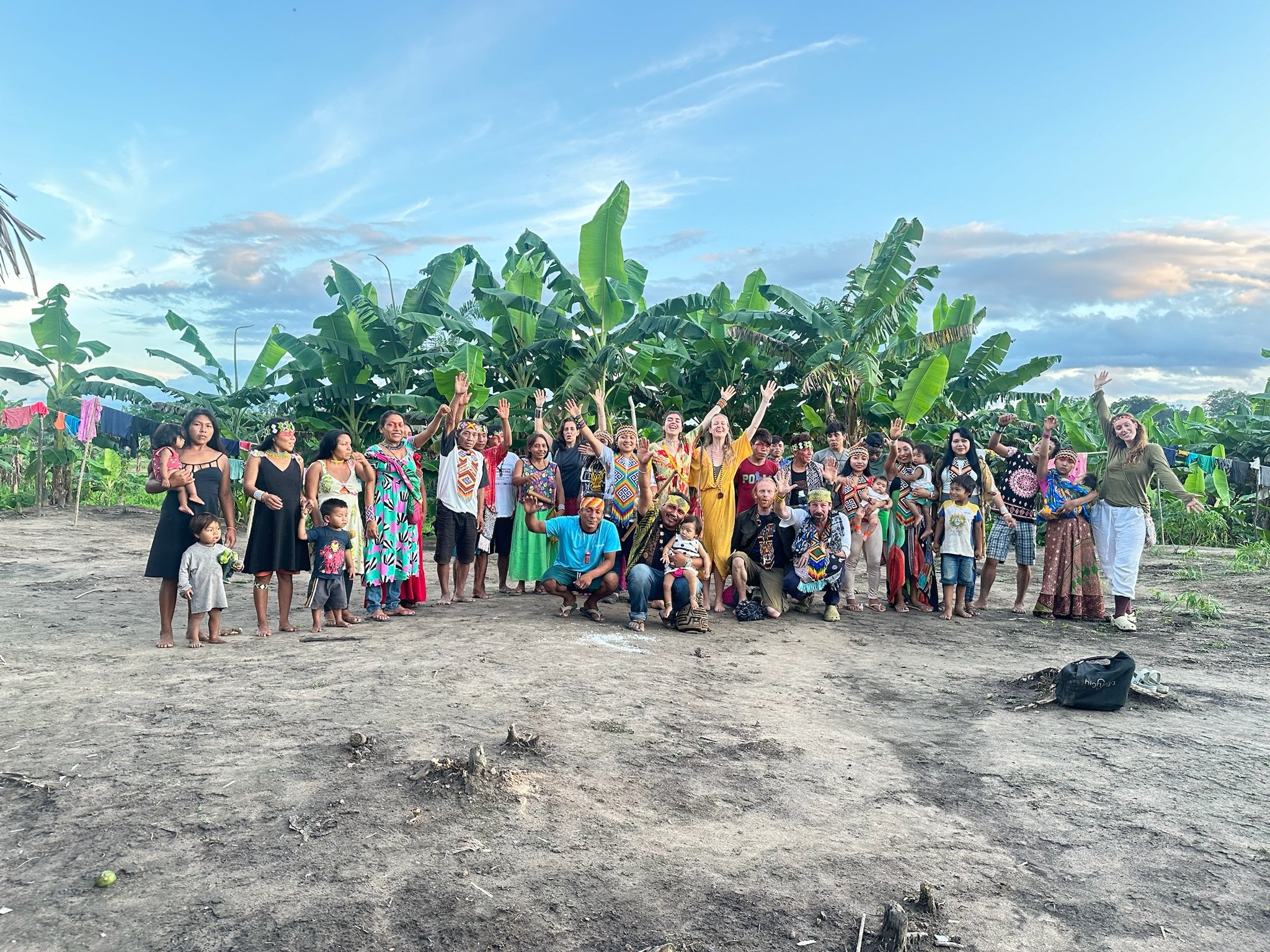 Grupo de pessoas indígenas posando em frente a uma plantação de bananeiras, com roupas tradicionais e modernas, sob céu parcialmente nublado.