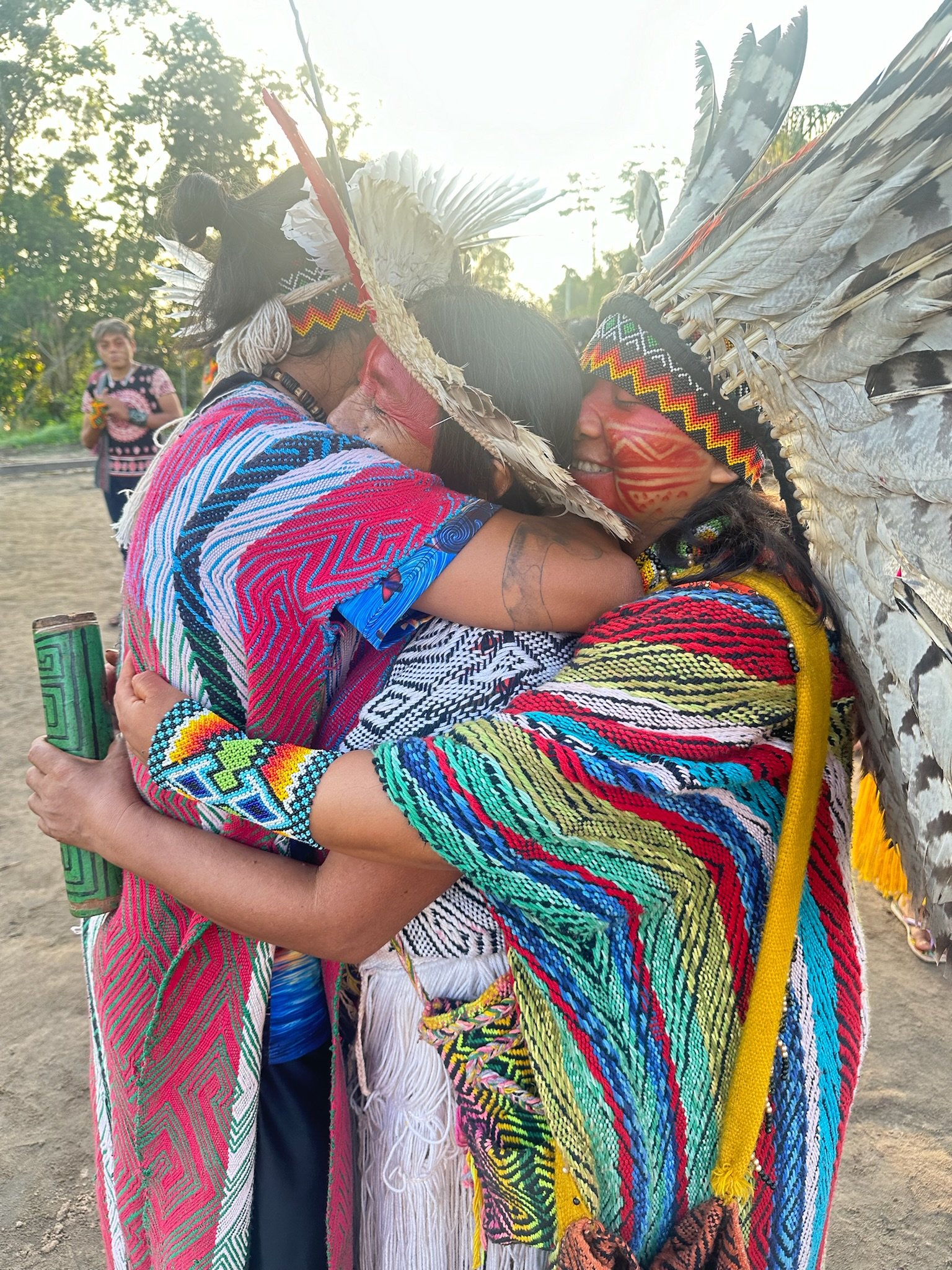Três pessoas indígenas em traje tradicional, estão abraçadas em um momento de celebração ao ar livre.