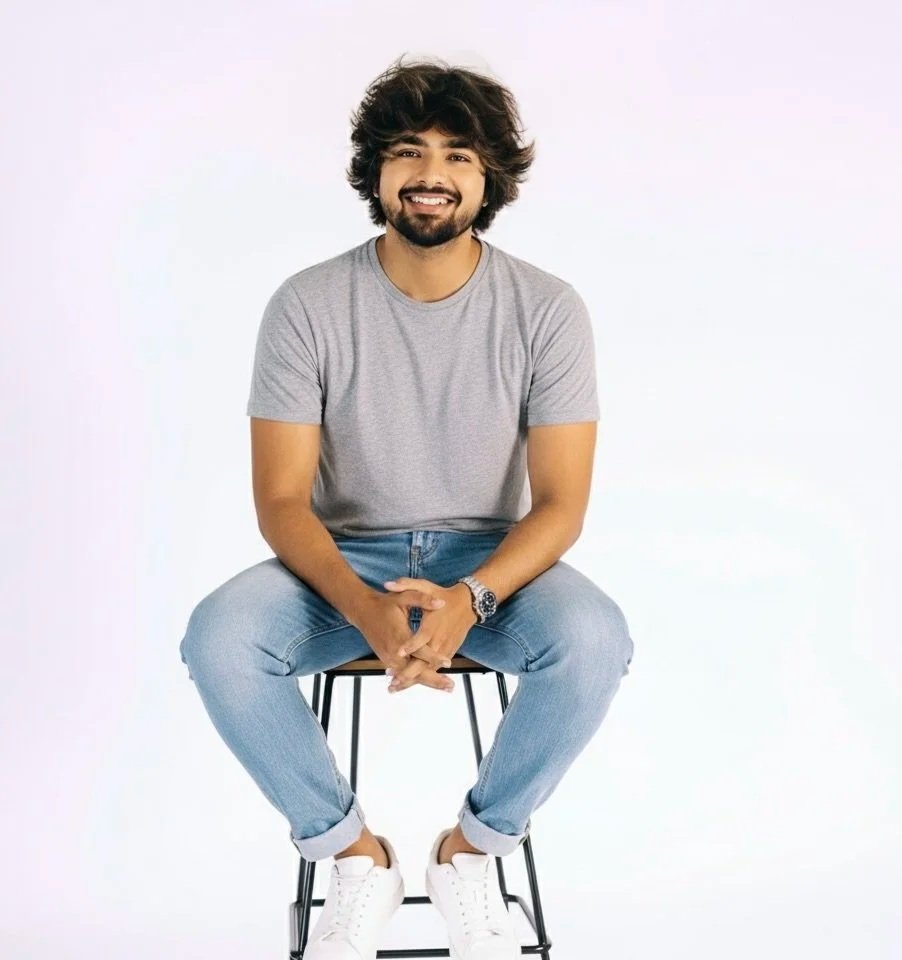A young man with curly dark hair and a beard sitting on a black stool, smiling, wearing a gray T-shirt, blue jeans, and white sneakers, with a light-colored plain background.