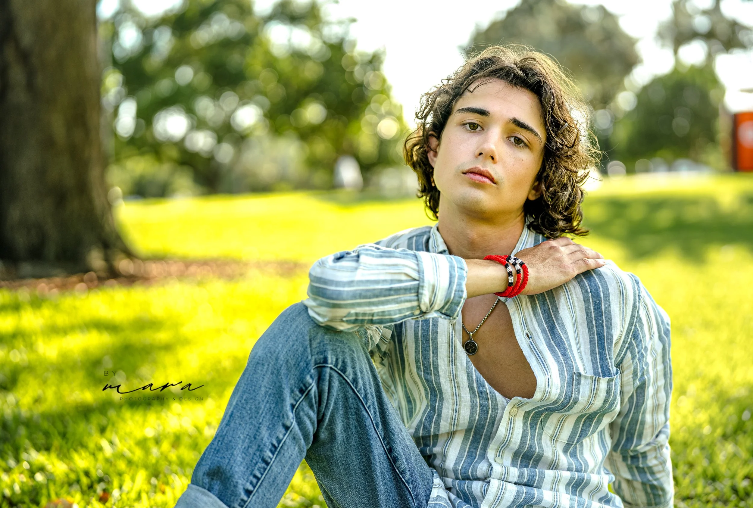 Person wearing a striped shirt and blue jeans sitting on grass in a park.