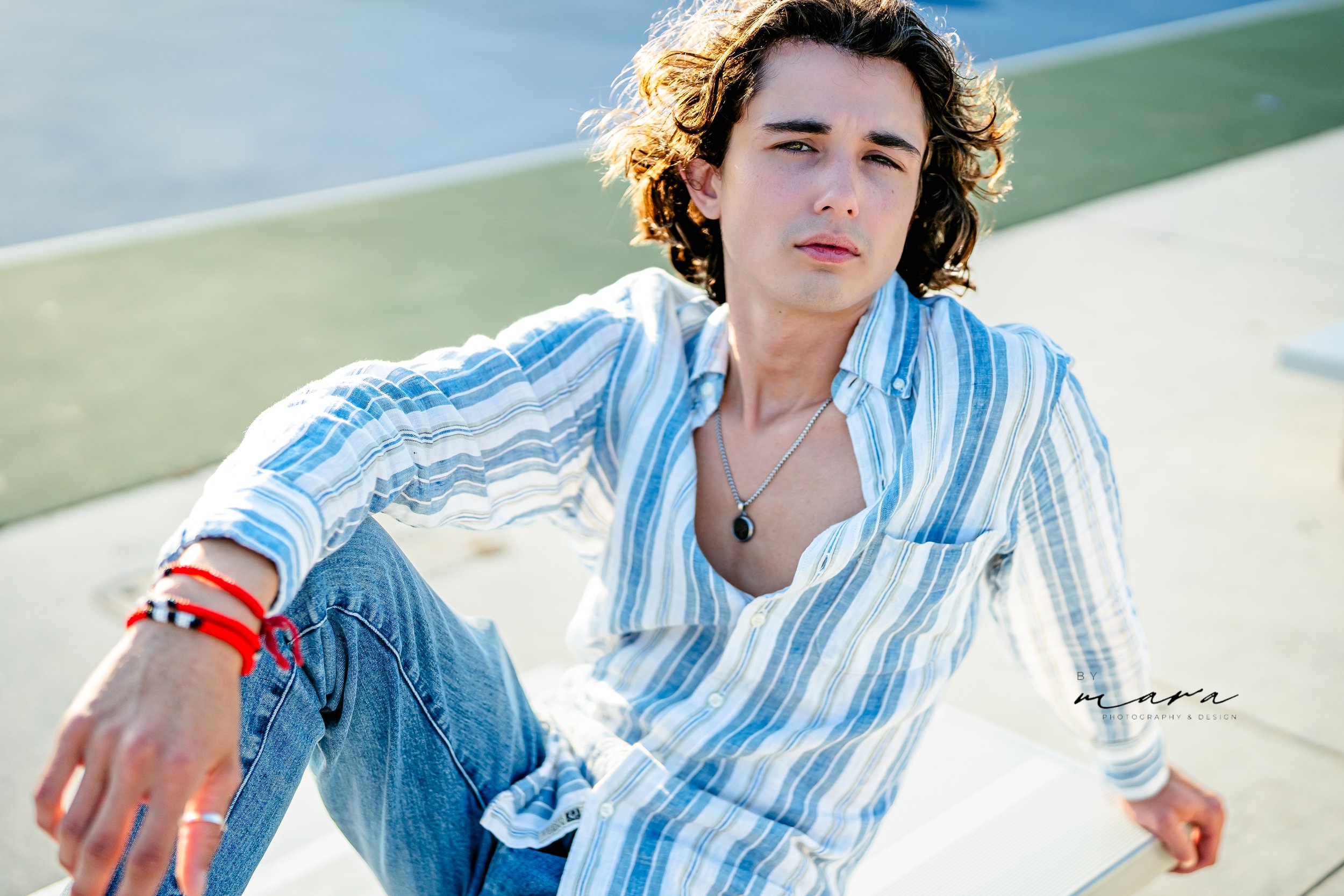 Person with wavy hair wearing a striped shirt and jeans, sitting outdoors on a sunny day.
