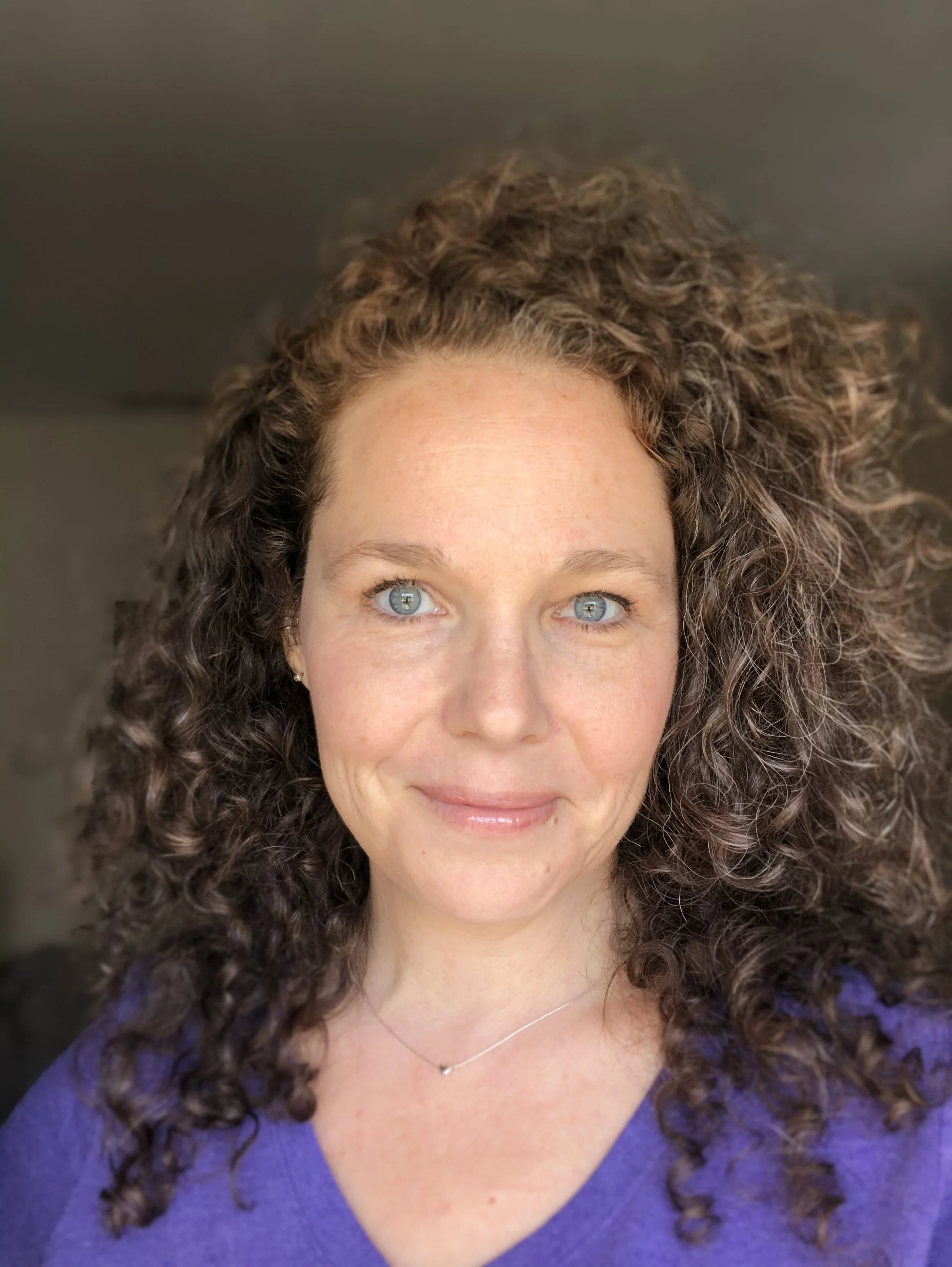 Person with curly hair wearing a purple shirt and a necklace, smiling slightly.