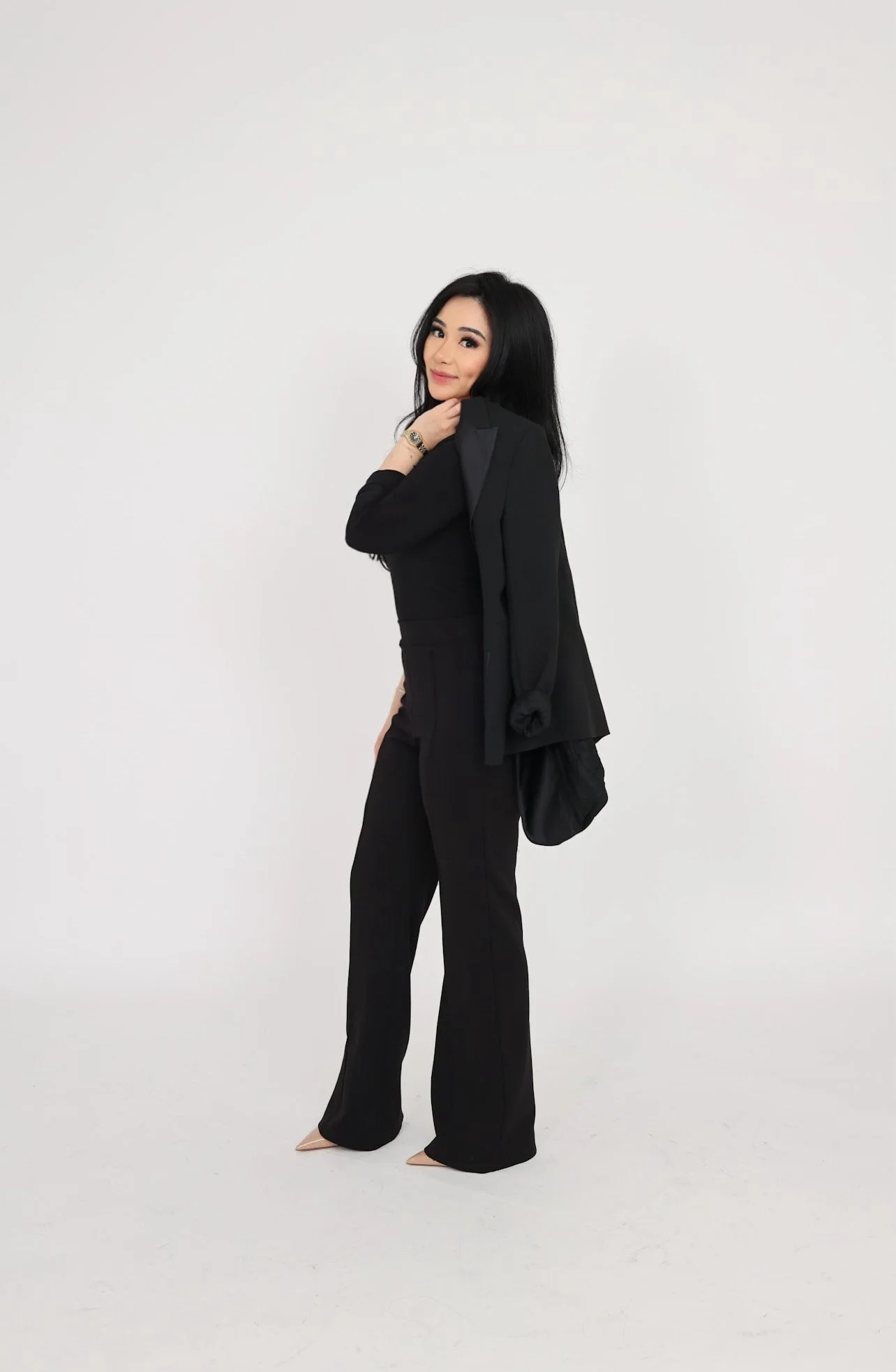 A woman with long black hair wearing a black outfit, smiling, standing against a plain white wall.