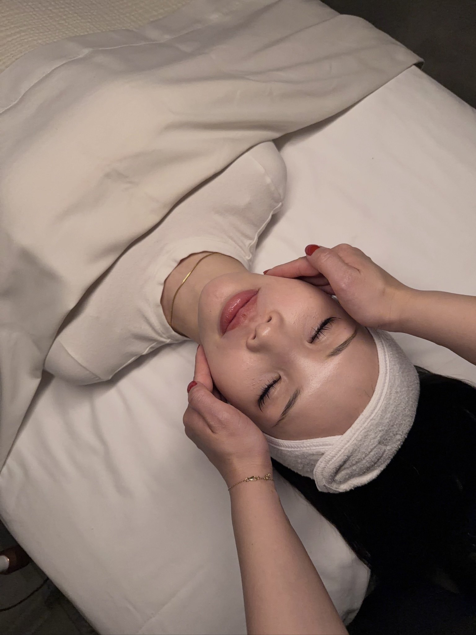 A woman receiving a facial massage at a spa, lying down with a white headband, eyes closed, and her face relaxed.