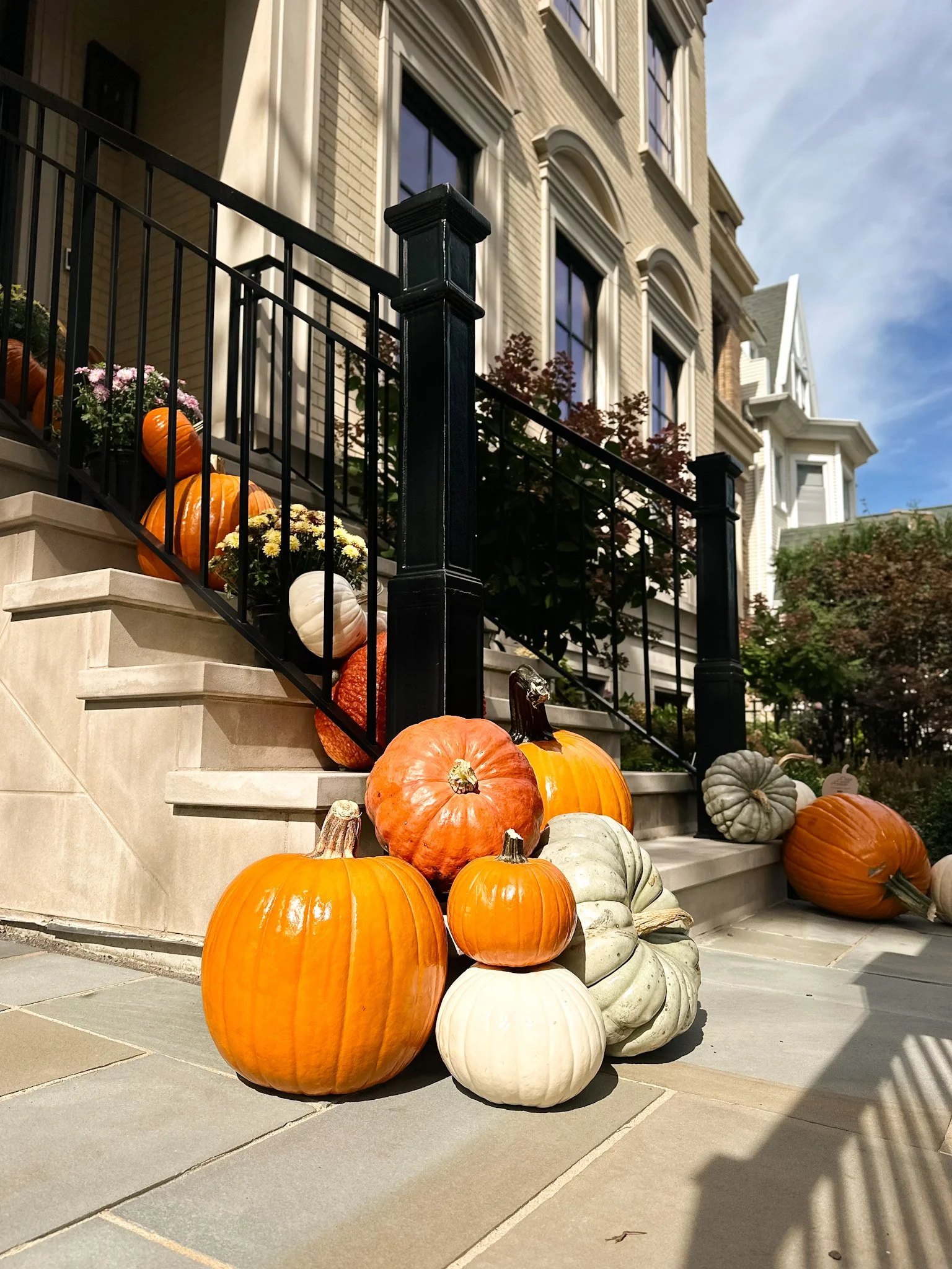 Front porch decorated with various pumpkins and gourds, flanking the stairs leading to a wooden front door with a wreath, black columns, and potted flowers on either side.