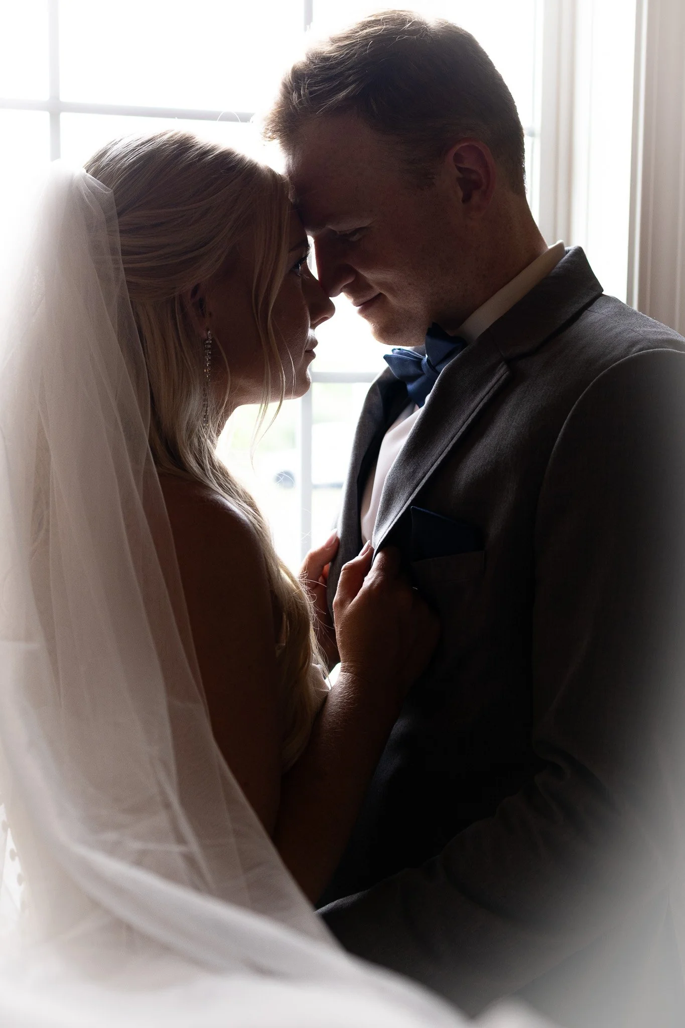 A bride and groom touching foreheads, standing close together indoors near a window, with backlit natural light behind them.
