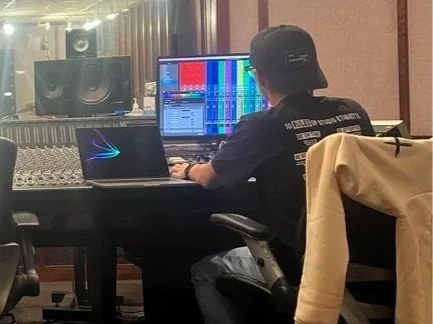 Person wearing a black cap and T-shirt working at a mixing console and computer in a recording studio.
