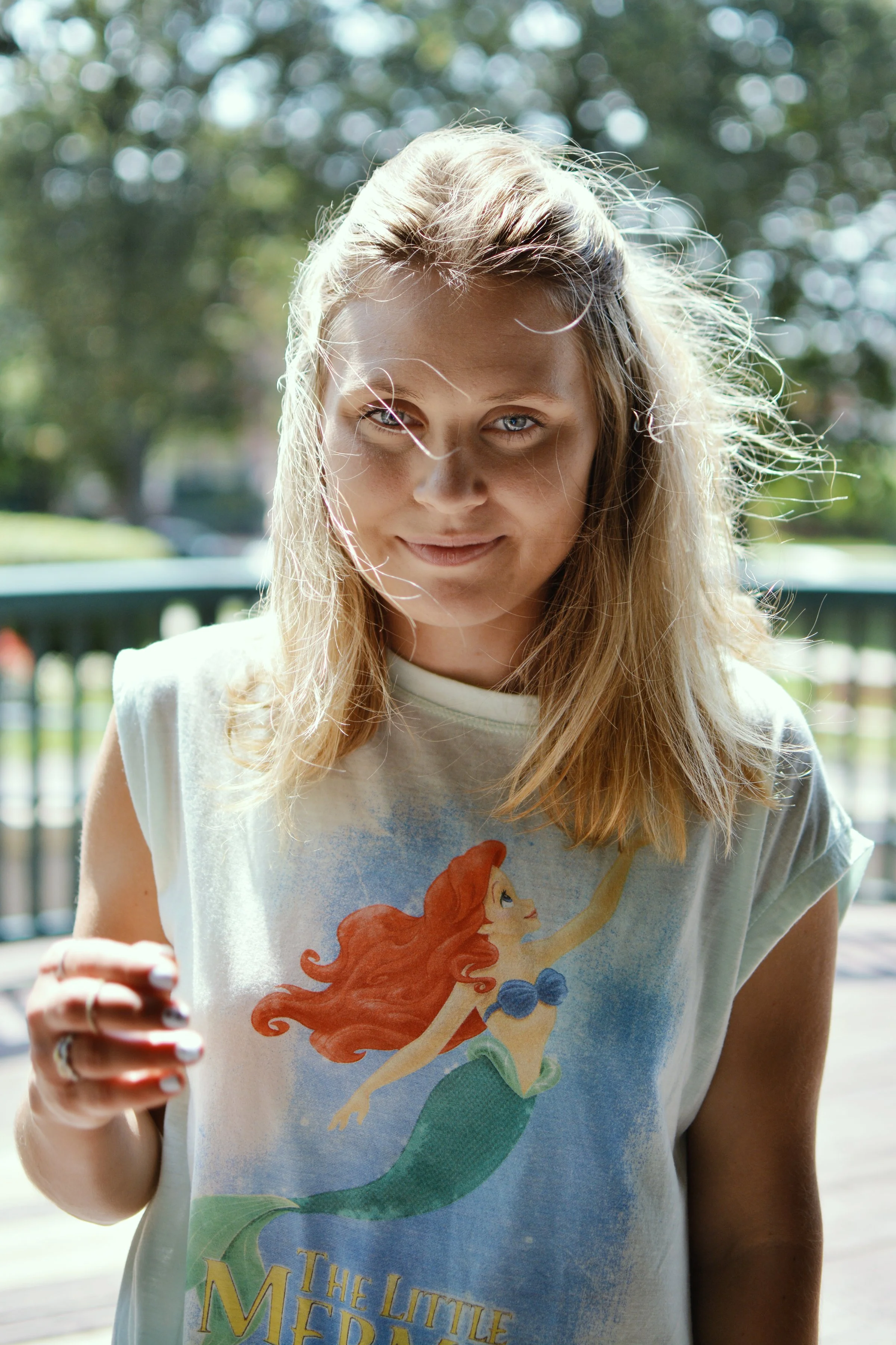 Close-up of a young woman with blonde hair and blue eyes, smiling slightly, wearing a white t-shirt with Ariel from Disney's The Little Mermaid on it, outdoors with blurred trees and a bench in the background.