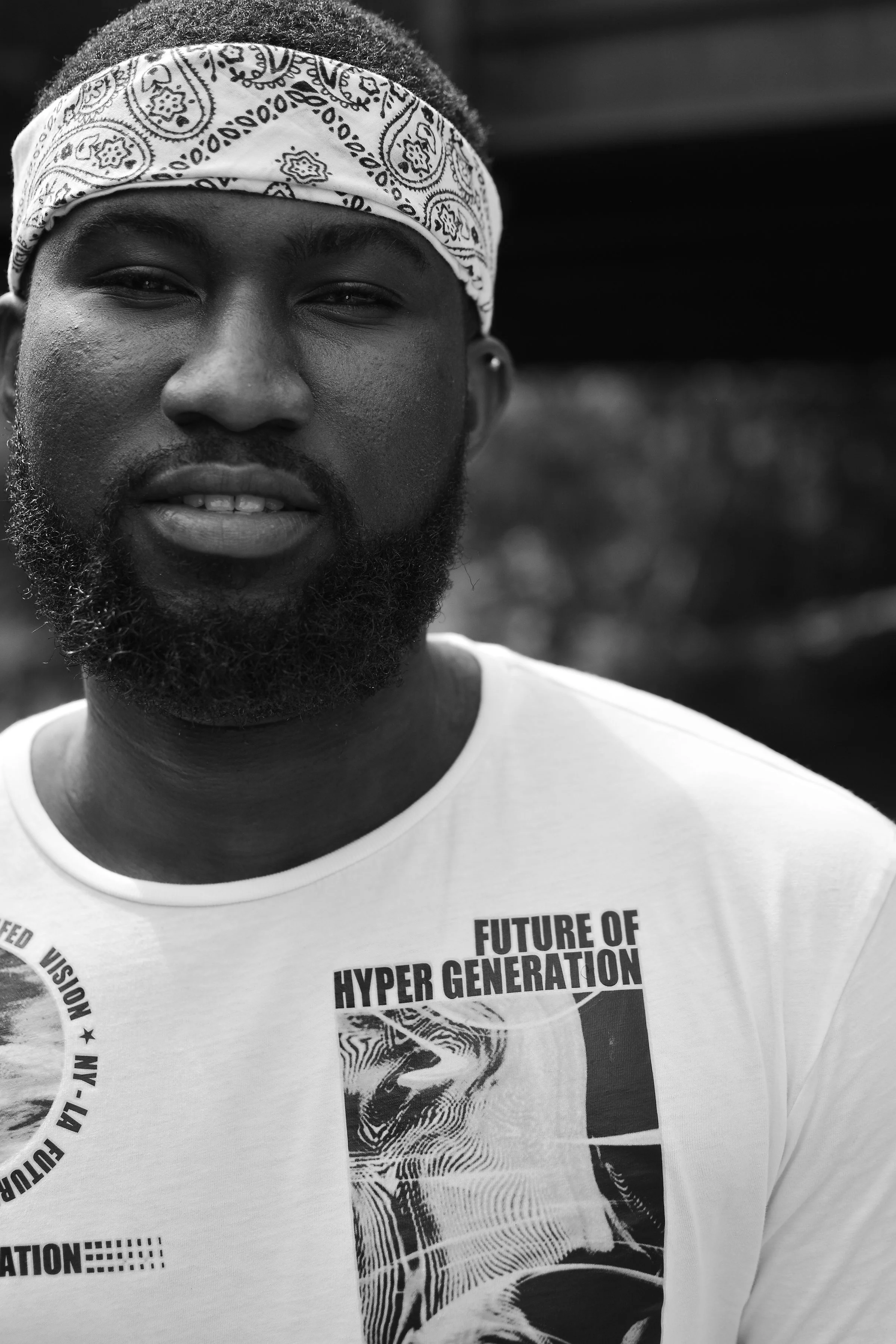 A young man with a beard, wearing a white bandana with paisley pattern and a white t-shirt with text and graphic designs, standing outdoors.