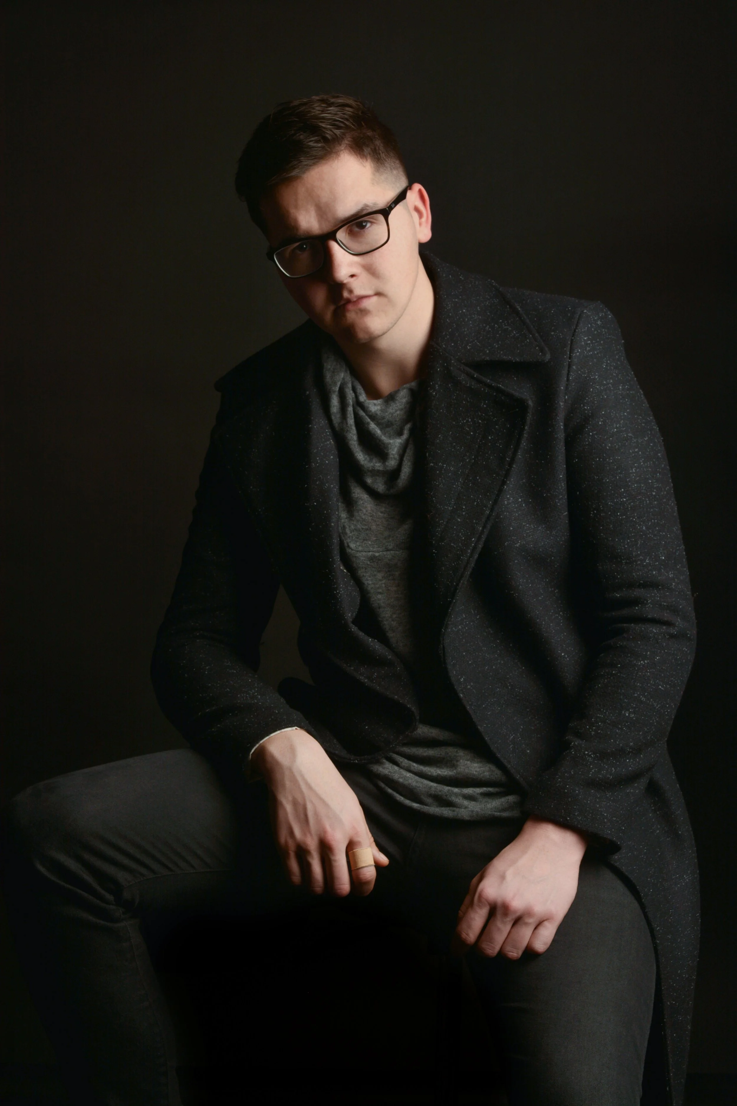 A young man with dark hair, wearing glasses, a speckled black coat, gray shirt, and black pants, sitting against a dark background with a serious expression.
