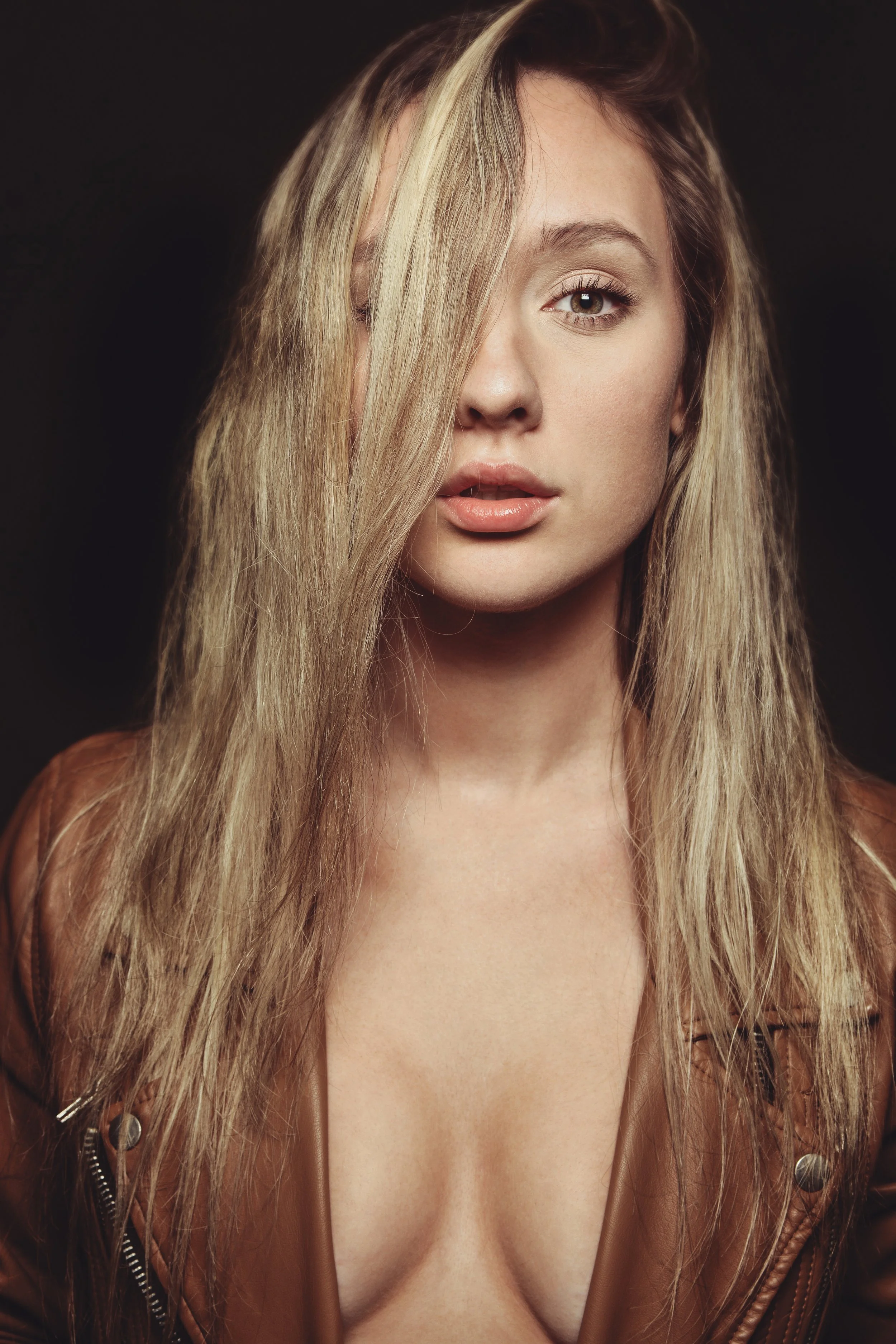 Close-up of a woman with long blonde hair covering one eye, wearing a brown leather jacket, with visible cleavage, against a dark background.