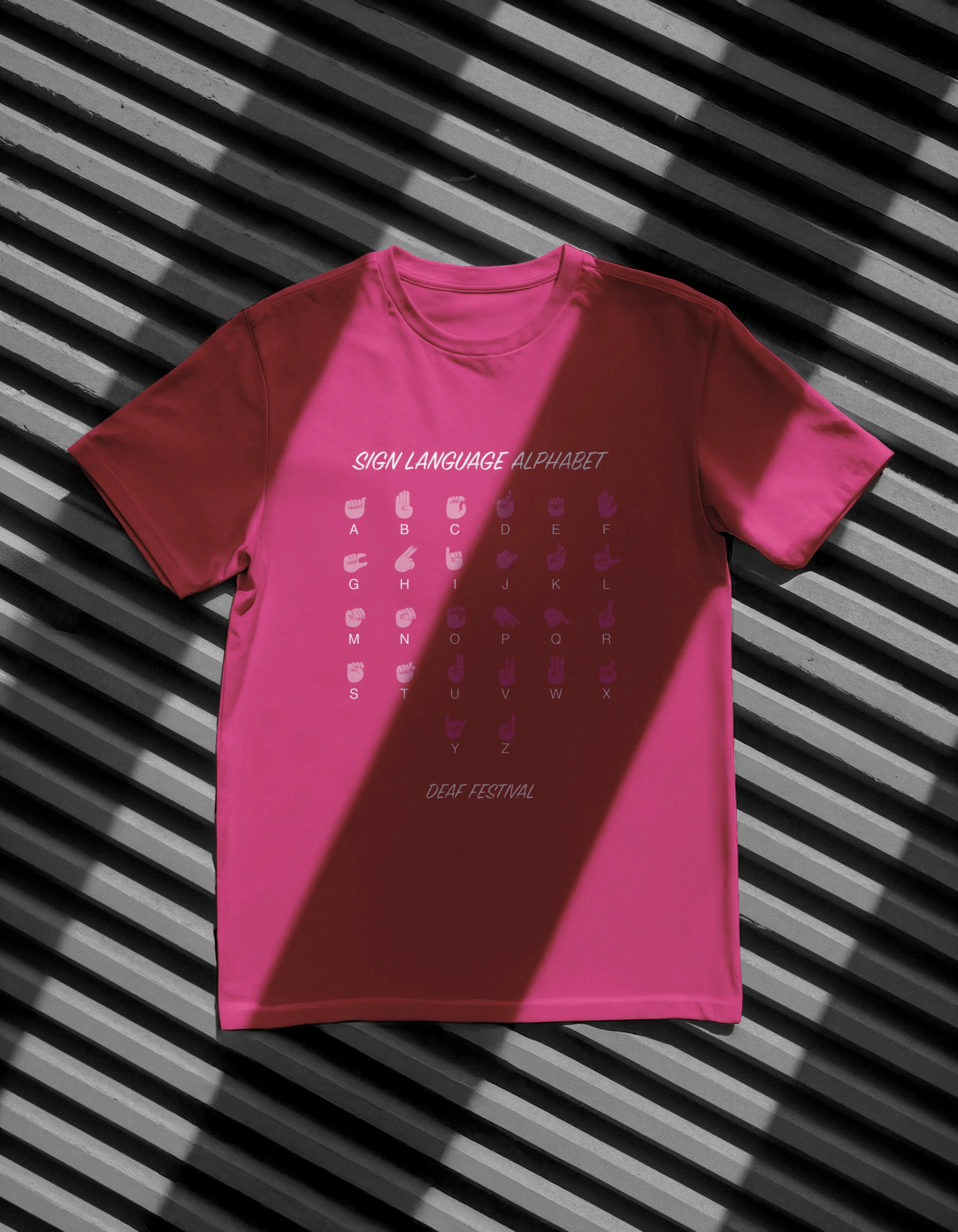Pink T-shirt with white text and sign language alphabet chart lying on a black and gray slatted surface.