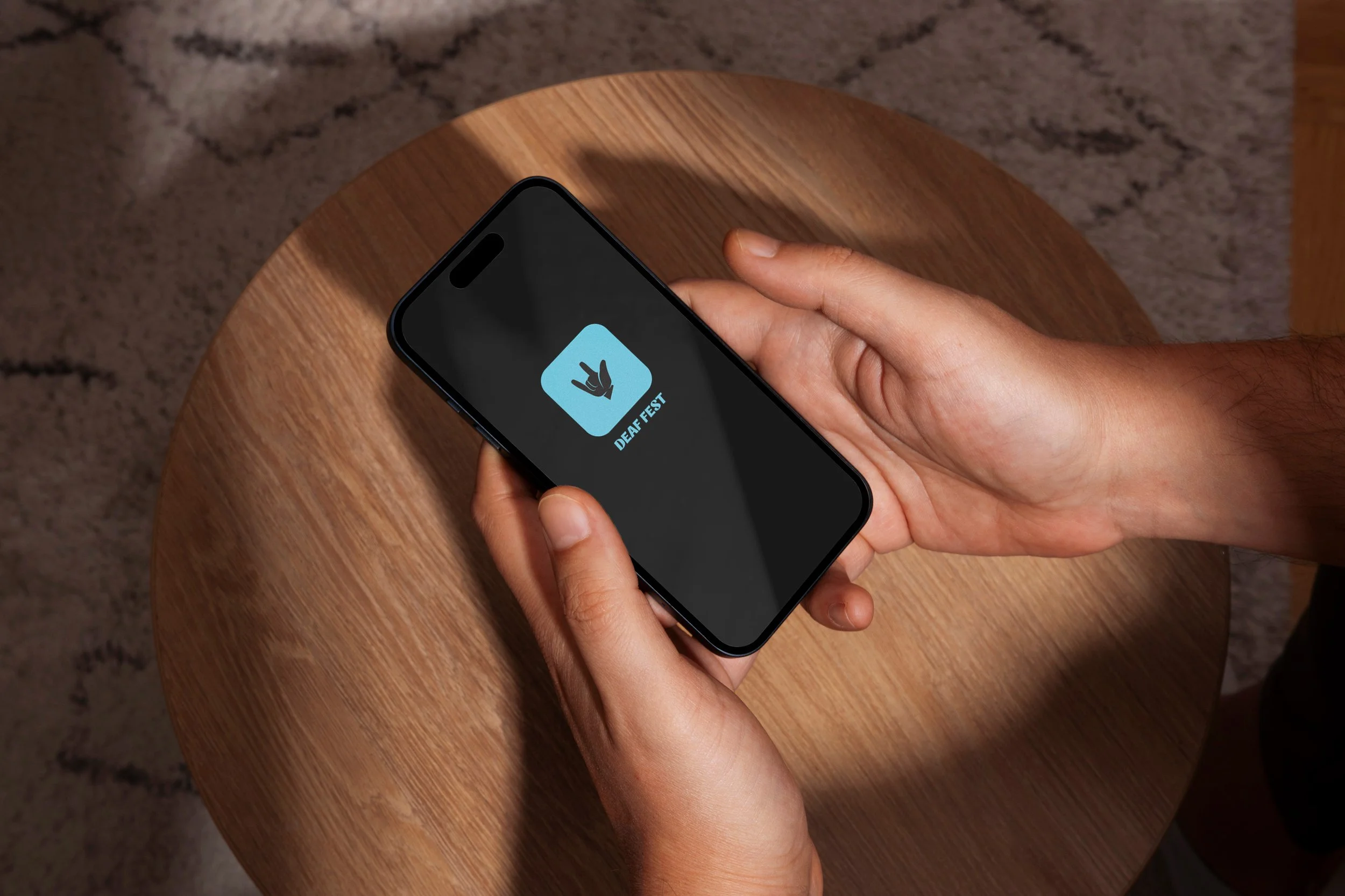 Person holding a smartphone with an app displaying the Deaf Fest logo, sitting at a wooden table.