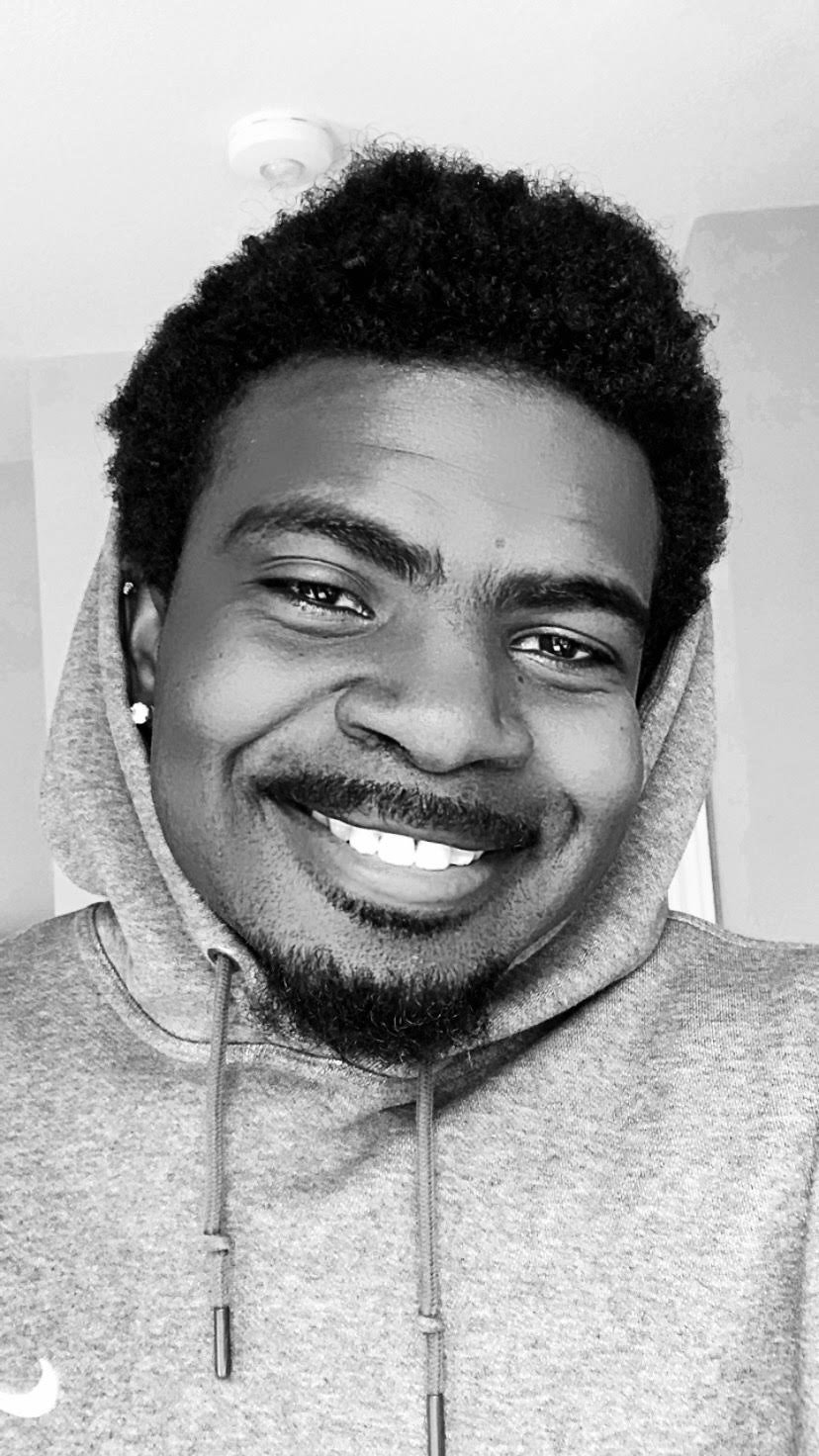 A smiling young man with a short curly beard, wearing a hoodie, earrings, and looking at the camera in a black and white photo.