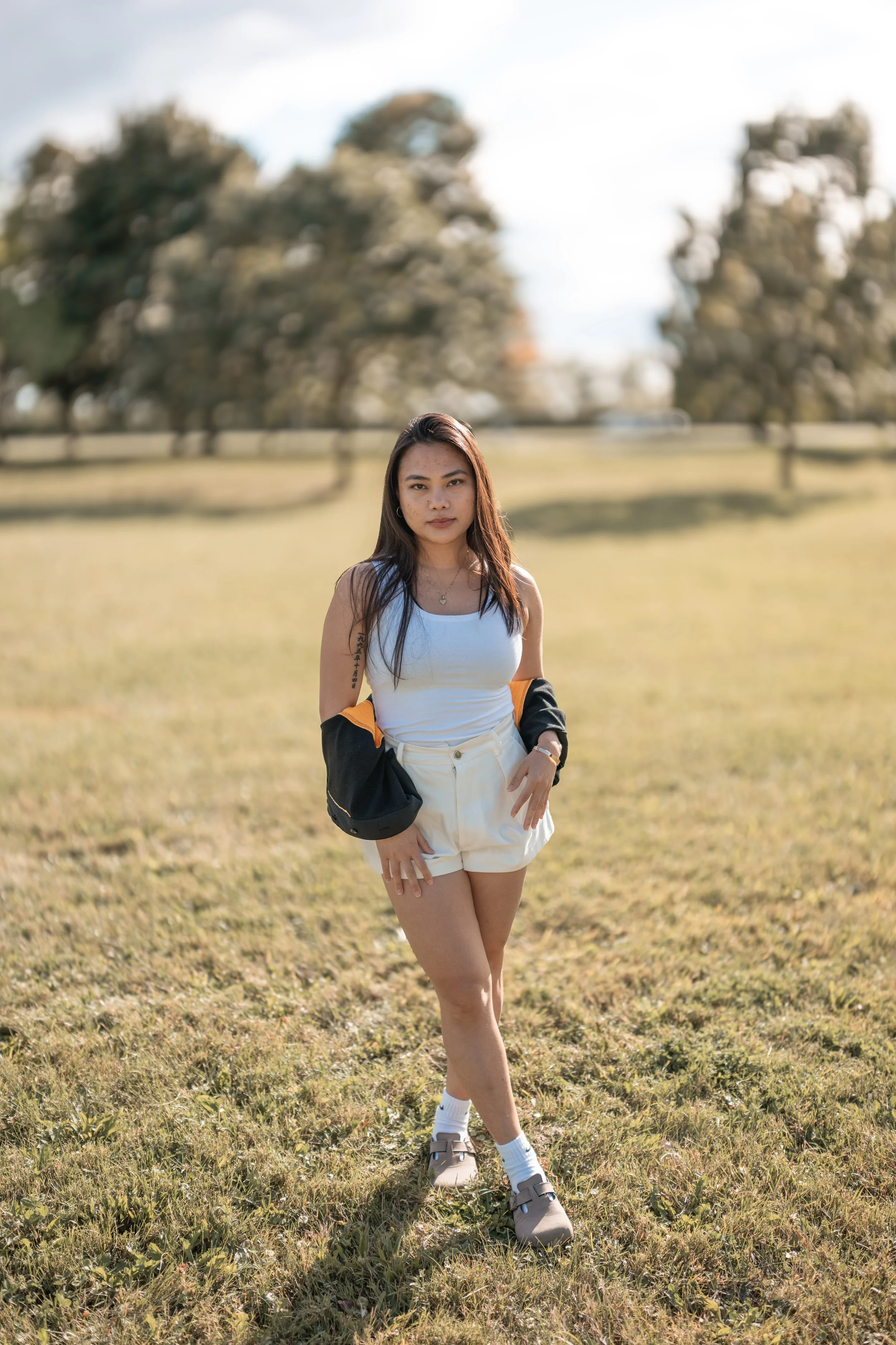 Young woman standing outdoors on a grassy field with trees and a fence in the background, looking at the camera with a neutral expression.