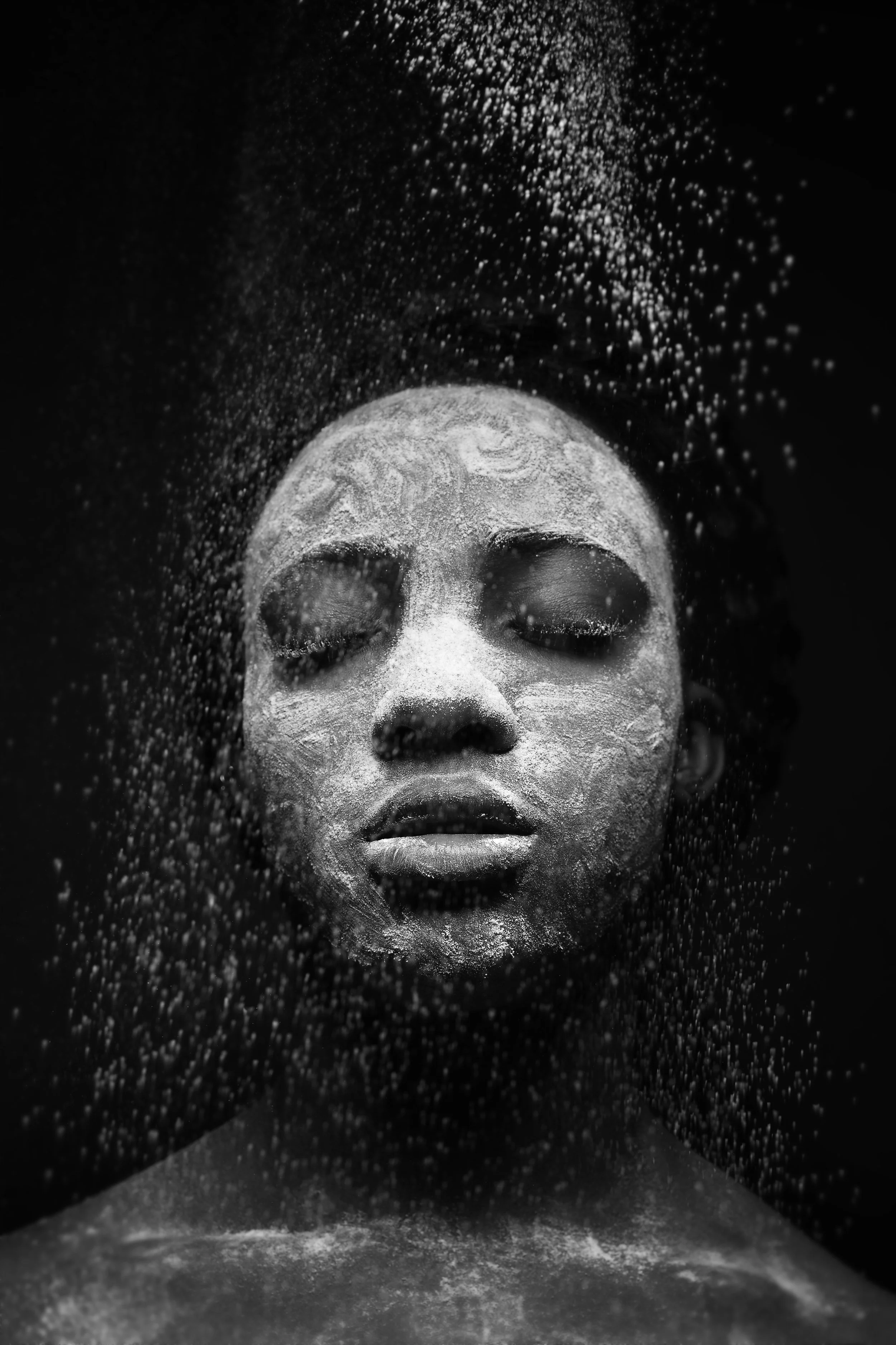 A woman with her eyes closed and face covered in a white facial mask. Powder is falling around her face, creating a dreamy, mystical atmosphere, against a dark background.