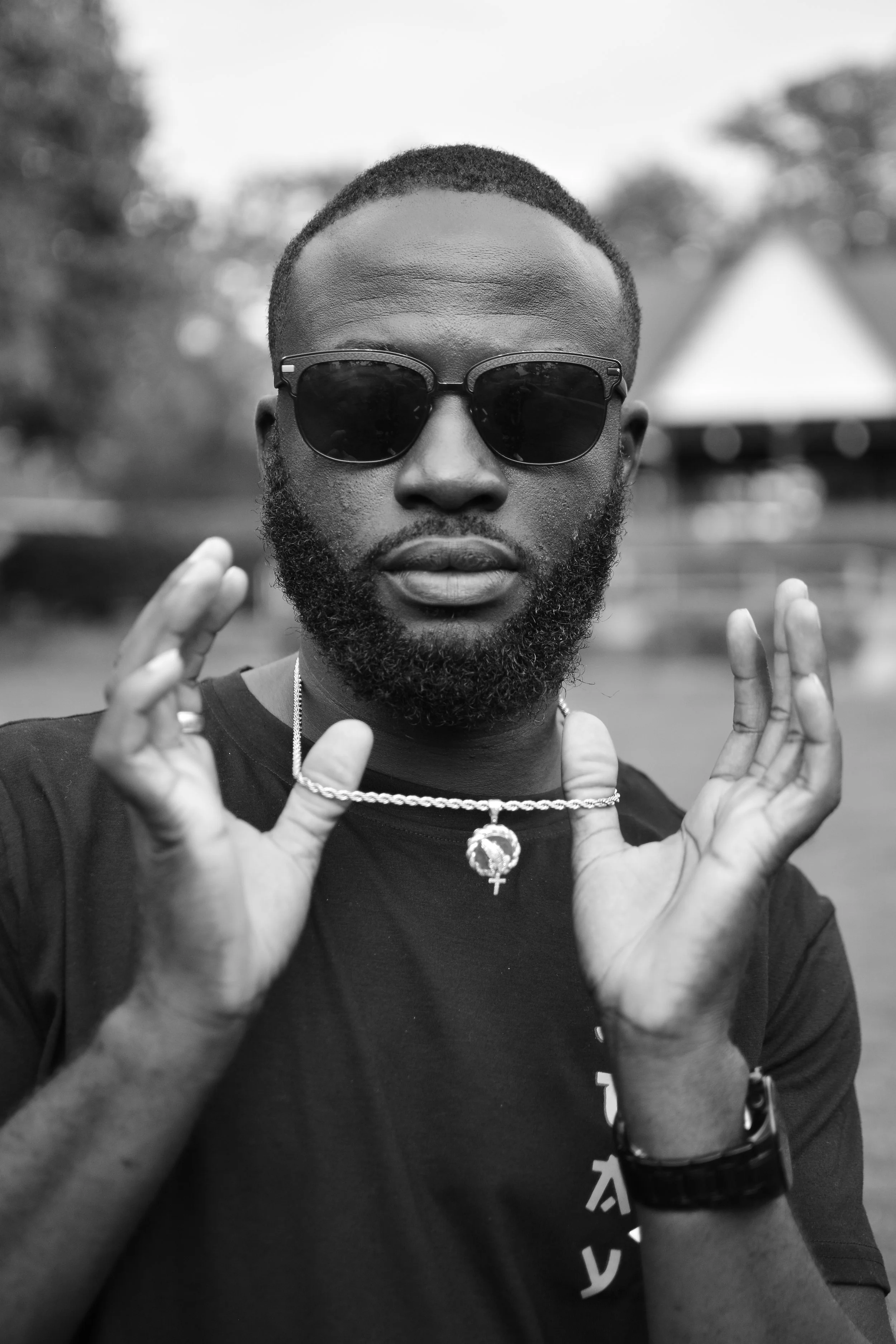 A black and white photo of a man wearing dark sunglasses, a necklace with a cross and peace sign pendant, and a black t-shirt. He has a beard and short hair, and is making a gesture with his hands near his face. The background appears to be outdoors 