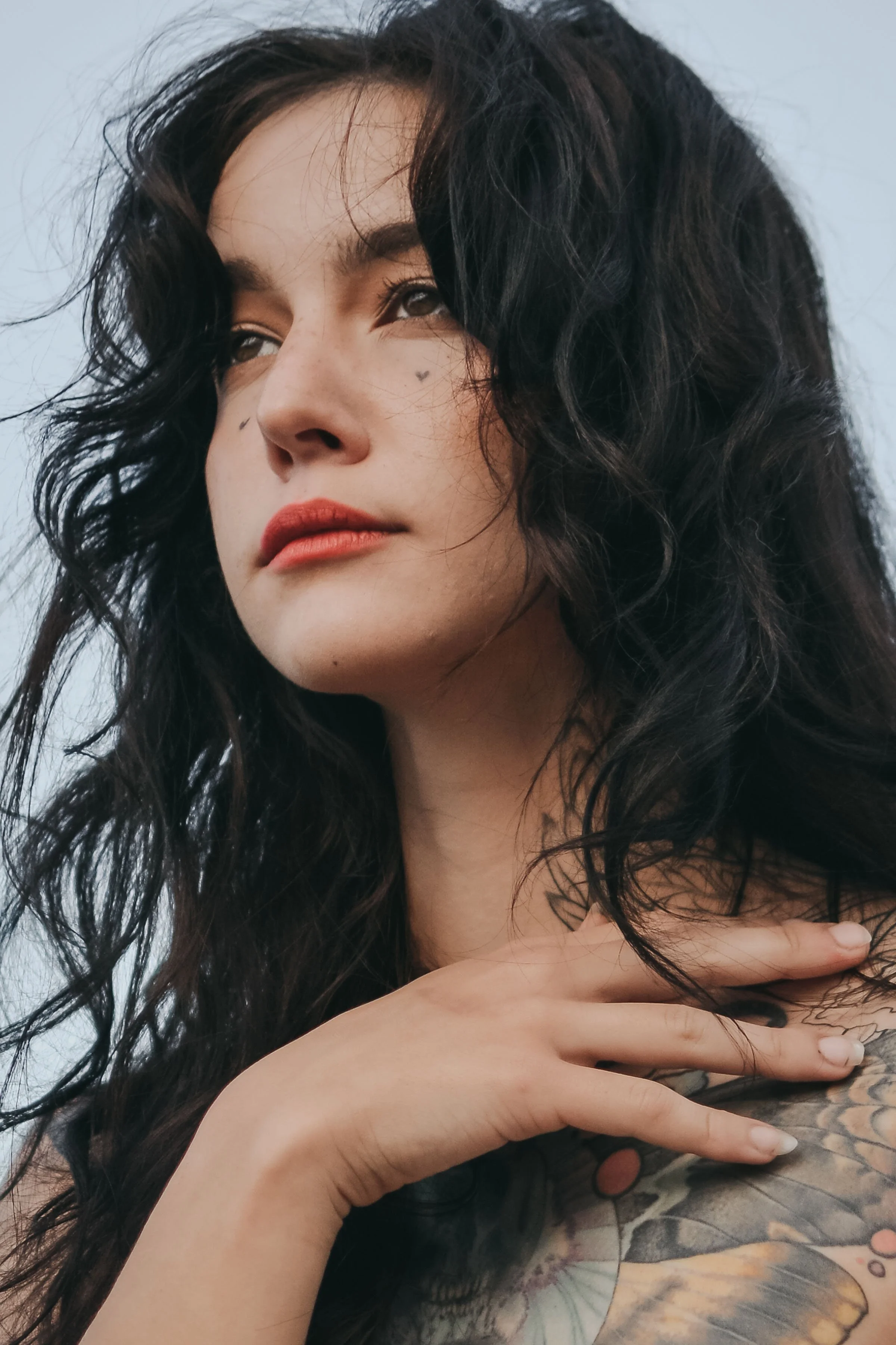 Close-up of a woman with dark curly hair, tattoos on her shoulder, and face tattoos of small heart and teardrop, looking thoughtfully to the side with hand on her shoulder.