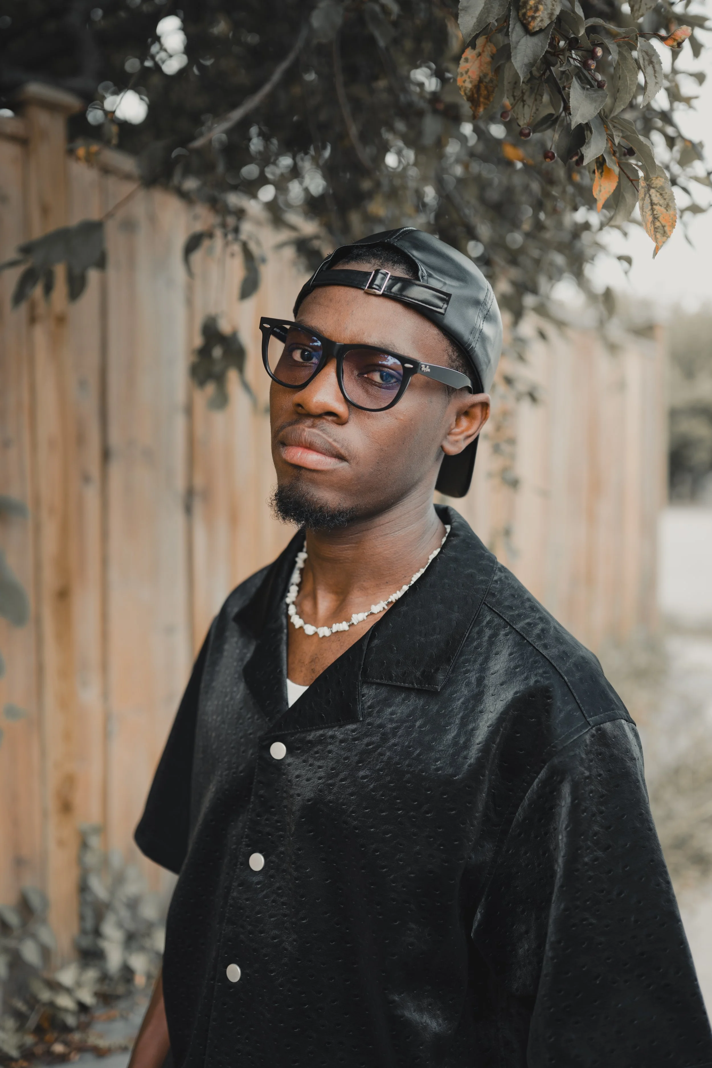 A young man wearing black sunglasses, a black leather cap worn backward, a black textured jacket with white buttons, and a pearl necklace poses outdoors in front of a wooden fence and leafy tree branches.