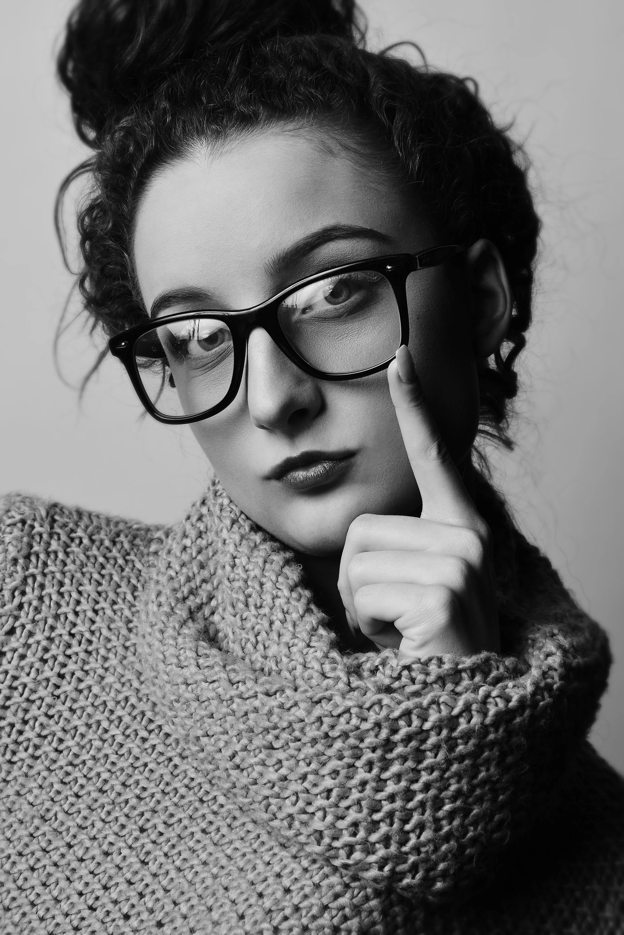 A woman wearing glasses and a knitted sweater, with curly hair, holding her finger to her lips in a contemplative pose in black and white.