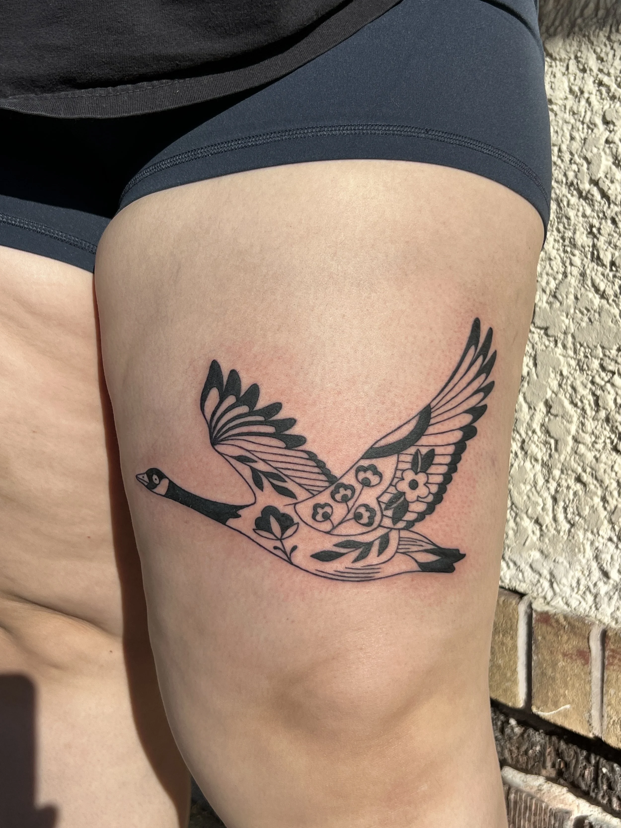 Close-up of a person's thigh with a black and gray tattoo of a bird with outstretched wings and flowers, wearing dark shorts and a black shirt, against a brick wall background.