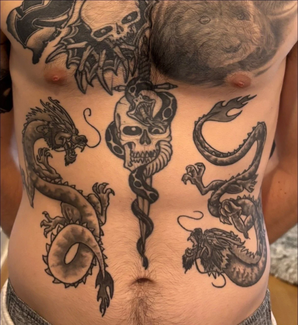 Tattooed male torso with a skull and snake in the center, two dragons on the sides, and a dog on the upper right chest.