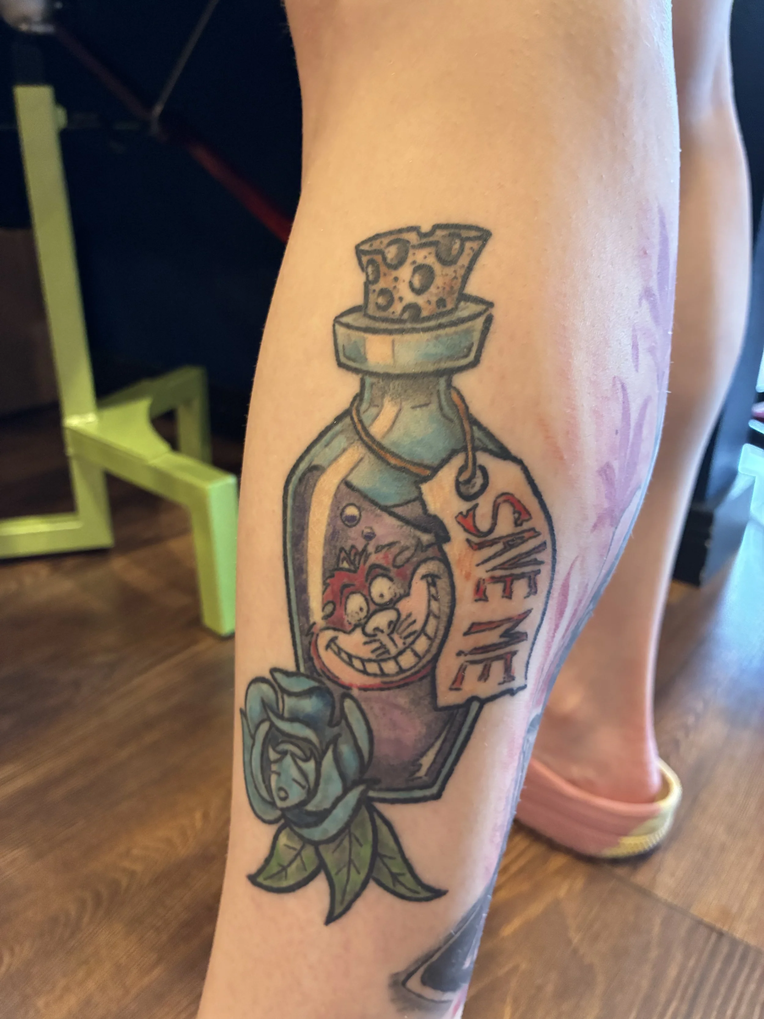 Tattoo of a cartoonish bottle with a smiling face and a pizza slice on top, surrounded by leaves.