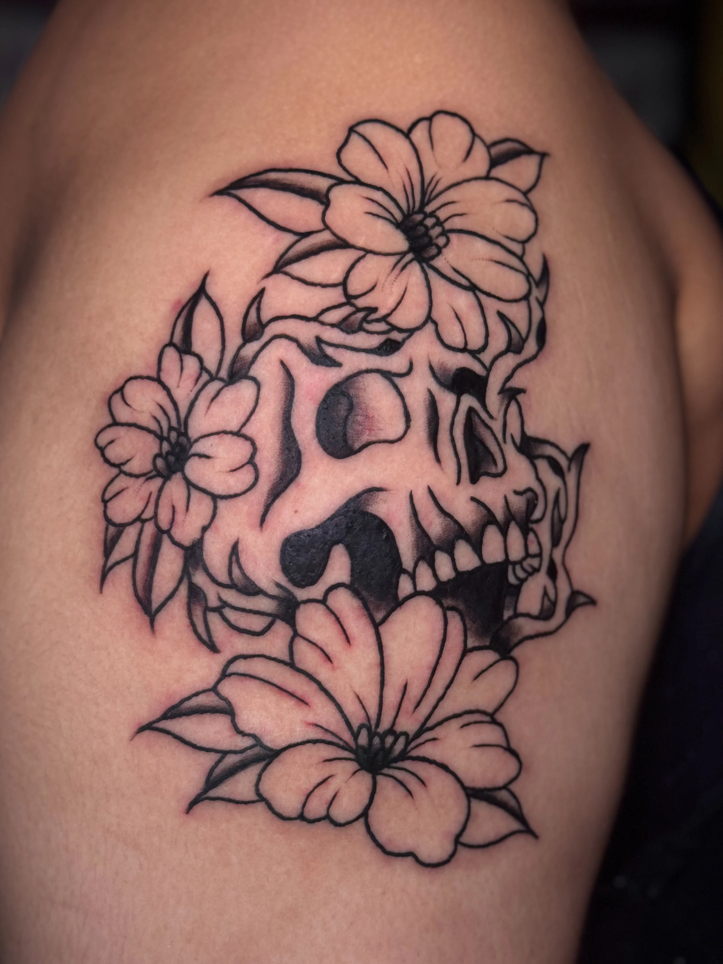 A close-up of a tattoo on skin featuring a detailed black outline of multiple flowers, including a large flower at the top with several leaves.