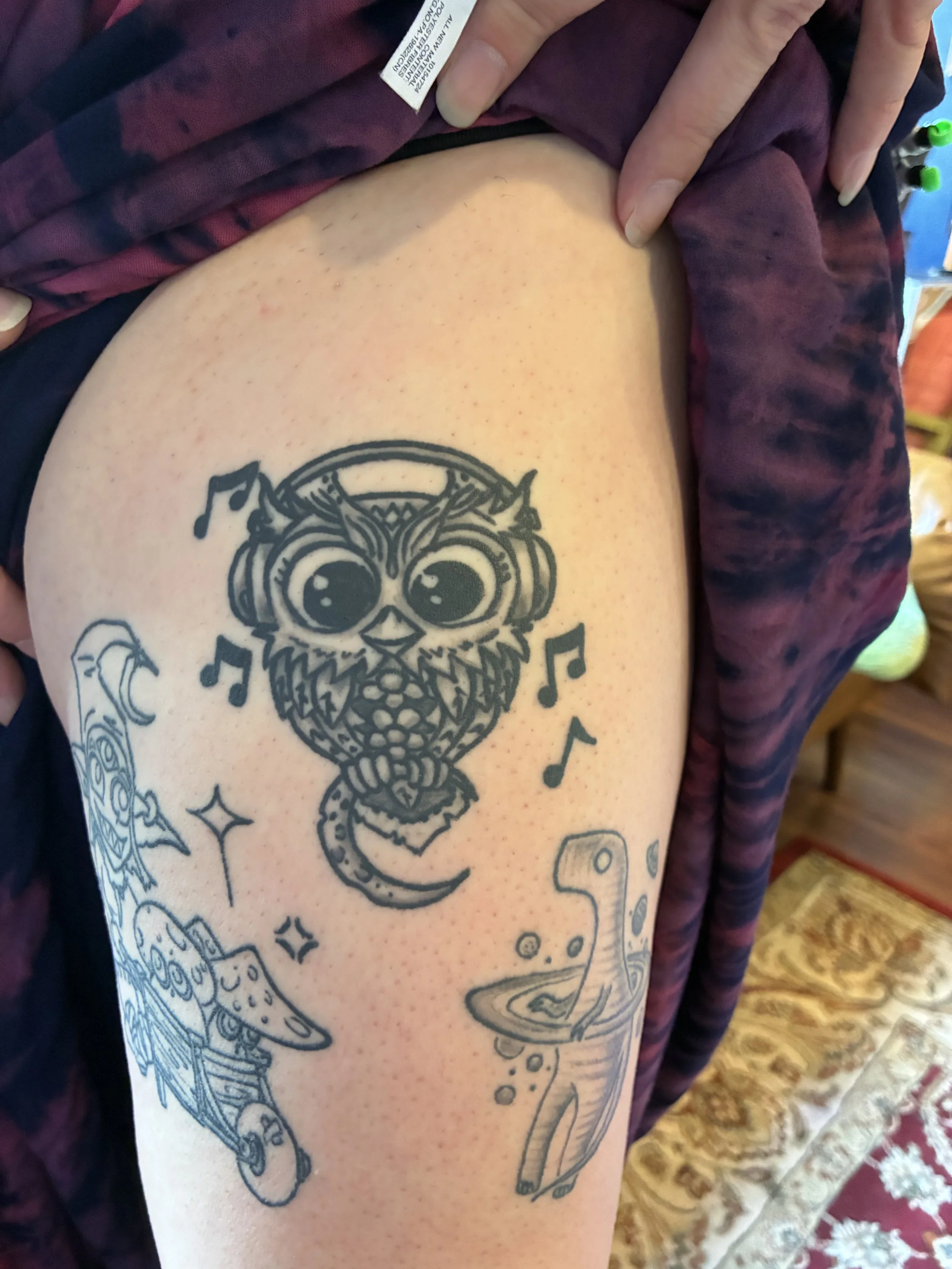 Tattoo on a person's upper arm featuring a cartoonish owl with musical notes surrounding it, a spaceship, and other decorative elements.