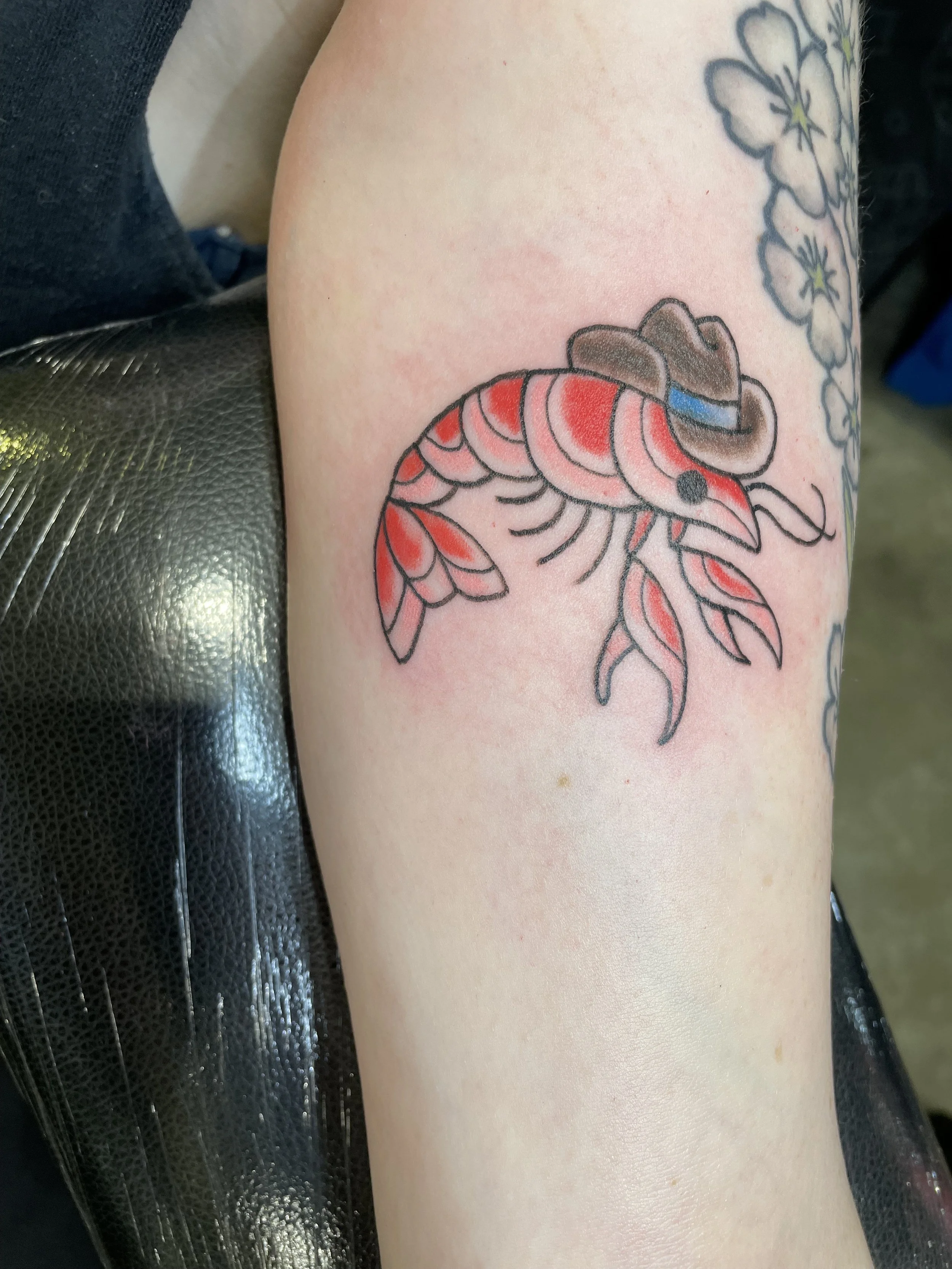 Tattoo of a stylized shrimp wearing a cowboy hat with a blue band, located on a person's upper arm.