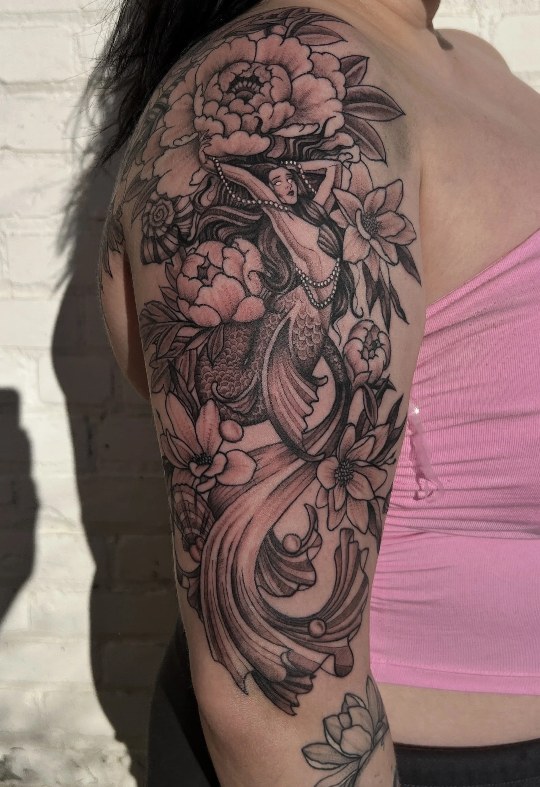 A detailed black and grey tattoo of a mermaid with long flowing hair, surrounded by large flowers and swirling patterns on a person's upper arm and shoulder.