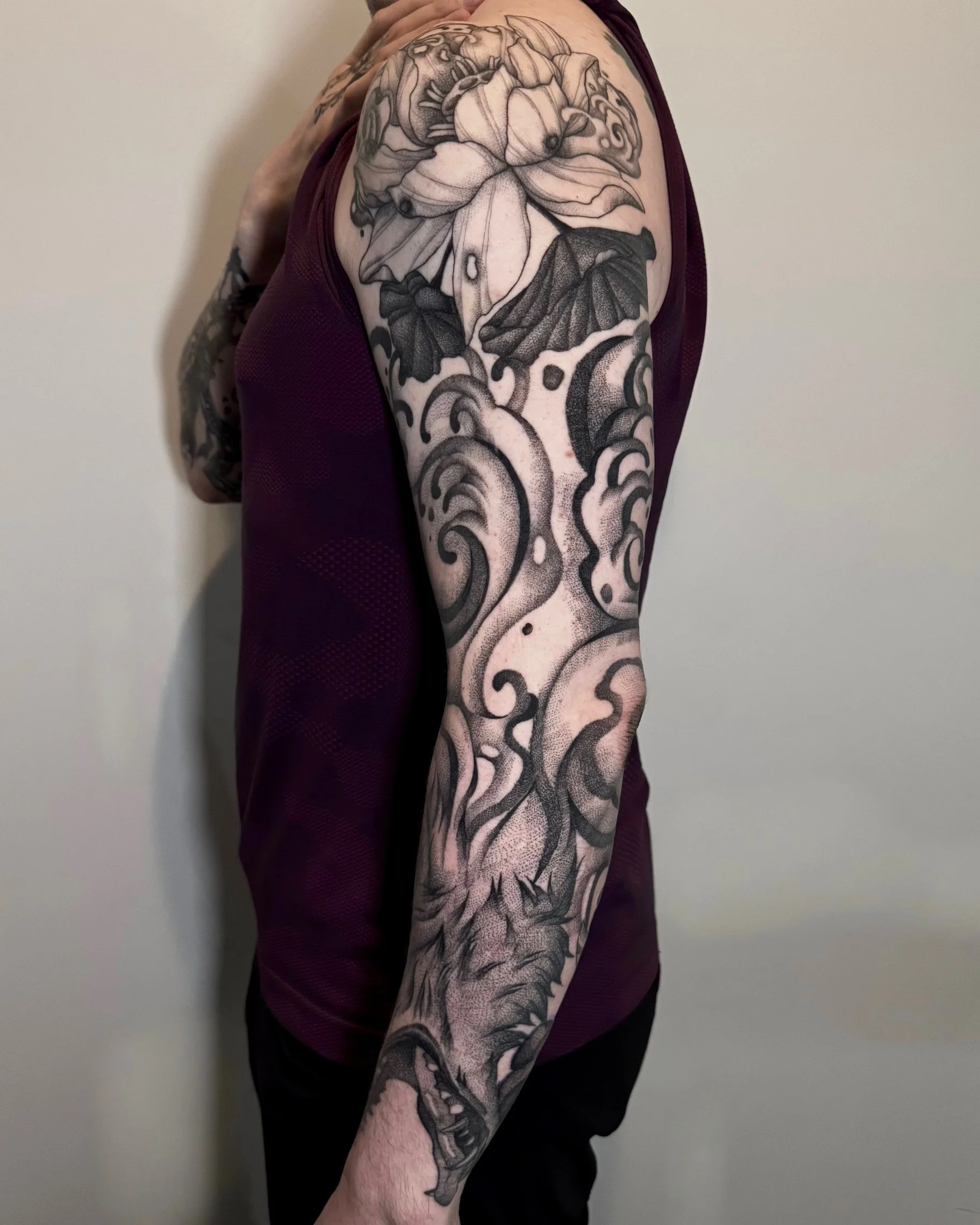 A person's upper arm and shoulder covered in a large black and gray tattoo featuring various floral and abstract designs.