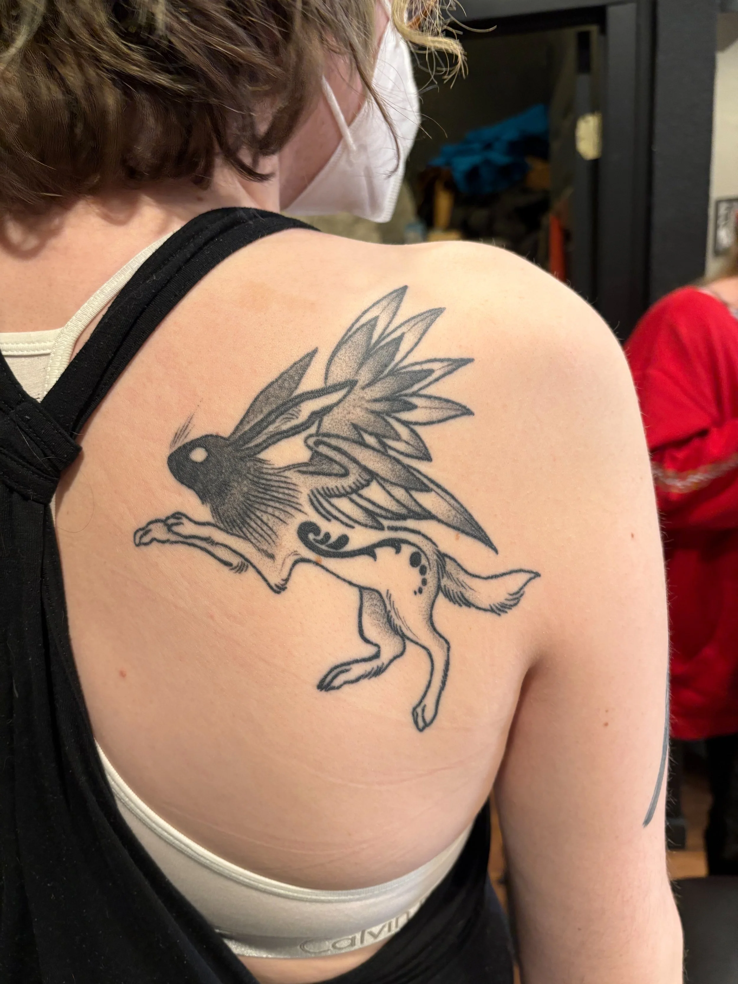 Tattoo of a mythical creature with the body of a wolf and the wings and head of a rabbit, drawn in black ink on a person's upper arm and shoulder.