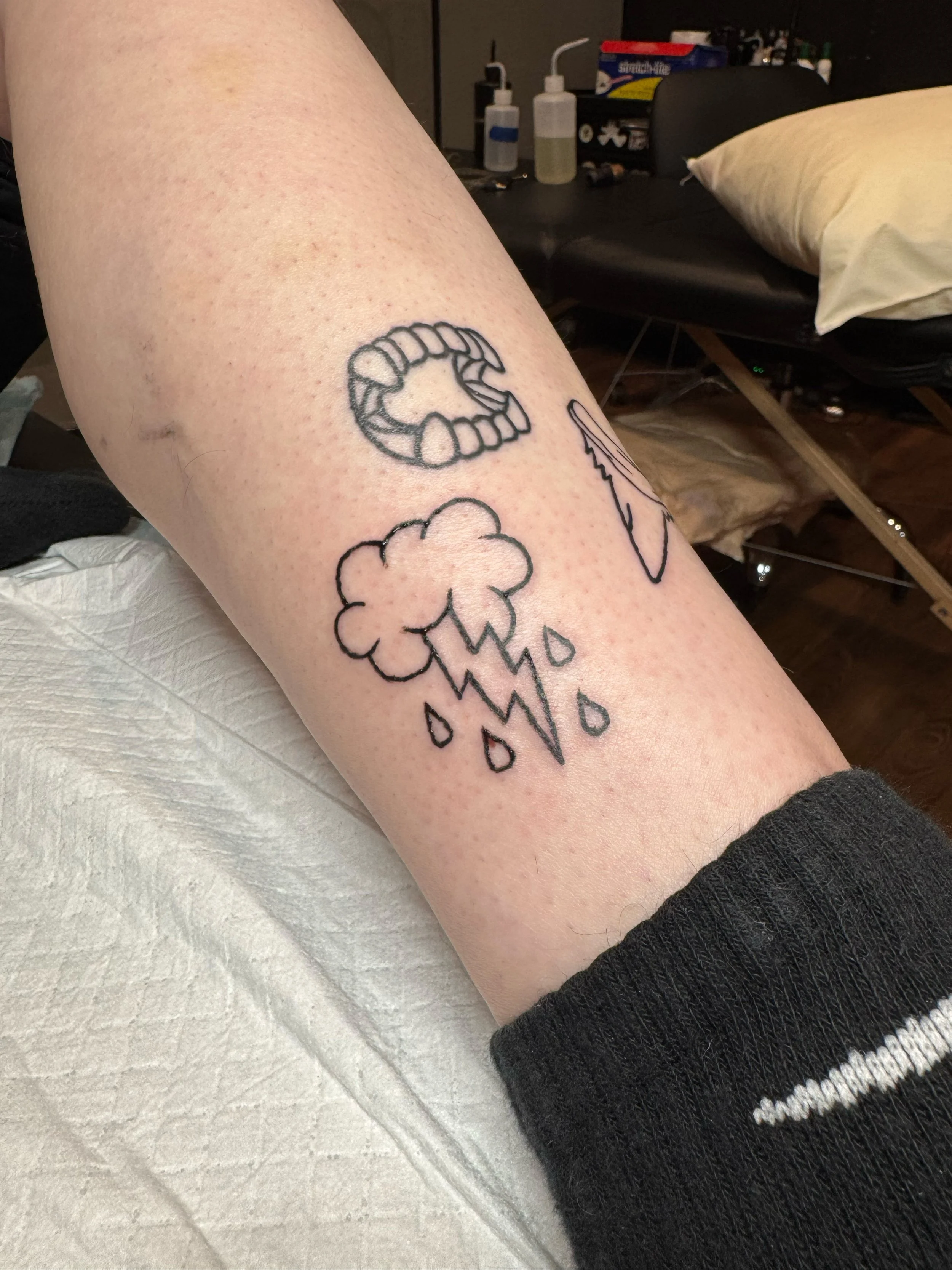 A tattoo on a person's forearm featuring a mouth with teeth, a cloud with rain drops, and a lightning bolt.
