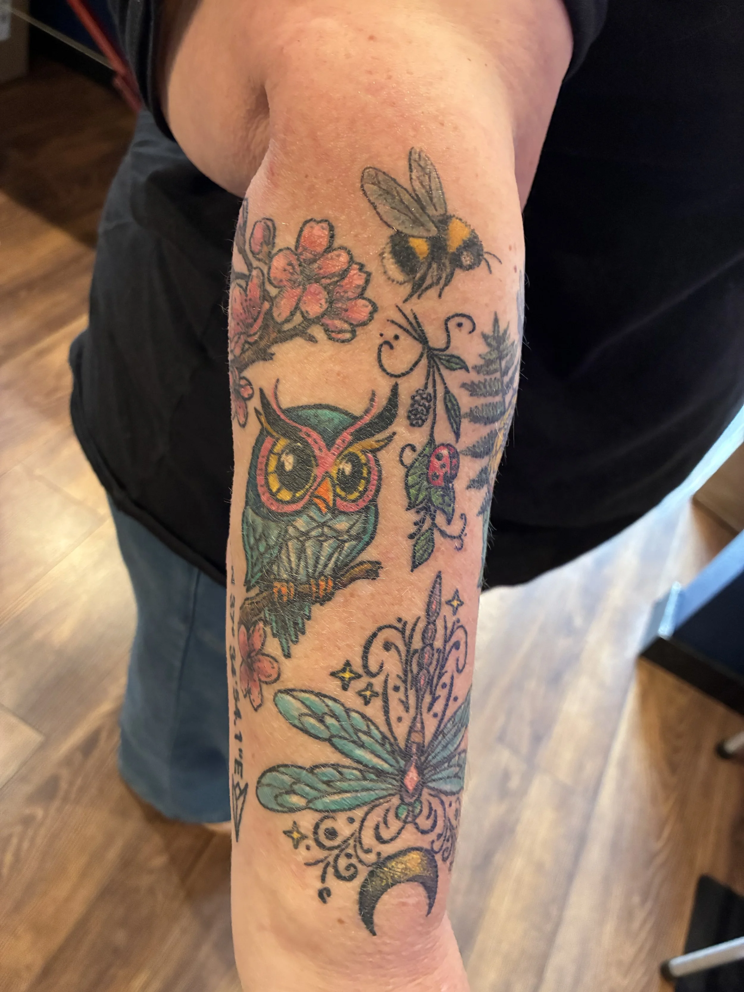 Tattooed arm with various colorful designs including a bee, butterfly, flower, and crescent moon.